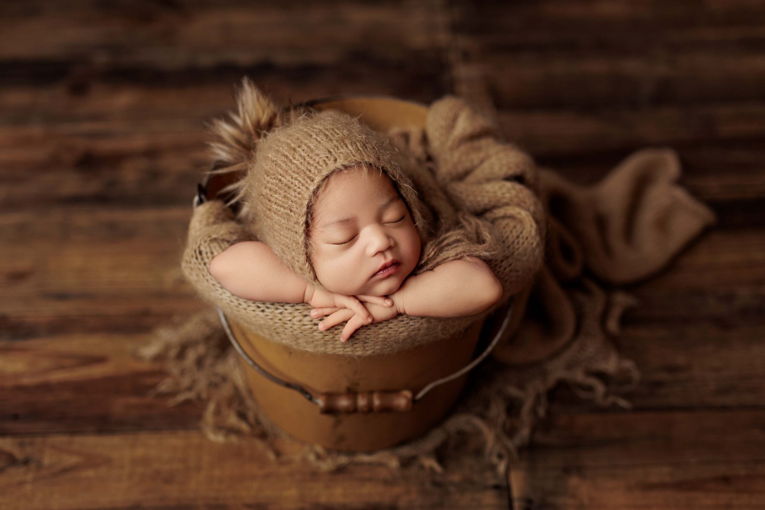 An Adelaide baby asleep in a tan bucket wearing a beanie with a fur pompom.