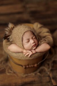 An Adelaide baby asleep in a tan bucket wearing a beanie with a fur pompom.