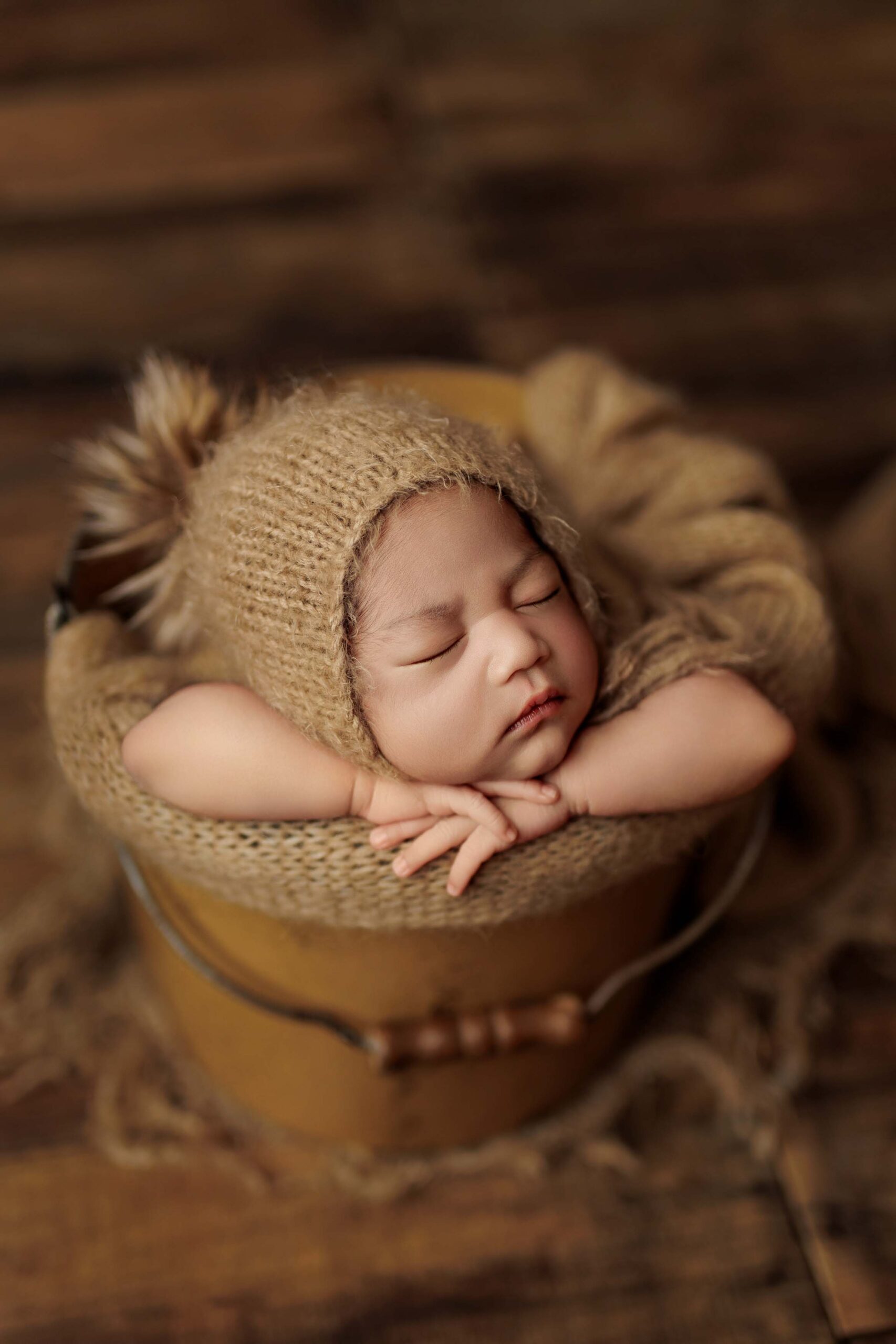 An Adelaide baby asleep in a tan bucket wearing a beanie with a fur pompom.