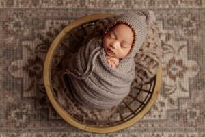An Adelaide newborn baby wrapped and wearing a bonnet asleep in a bowl.