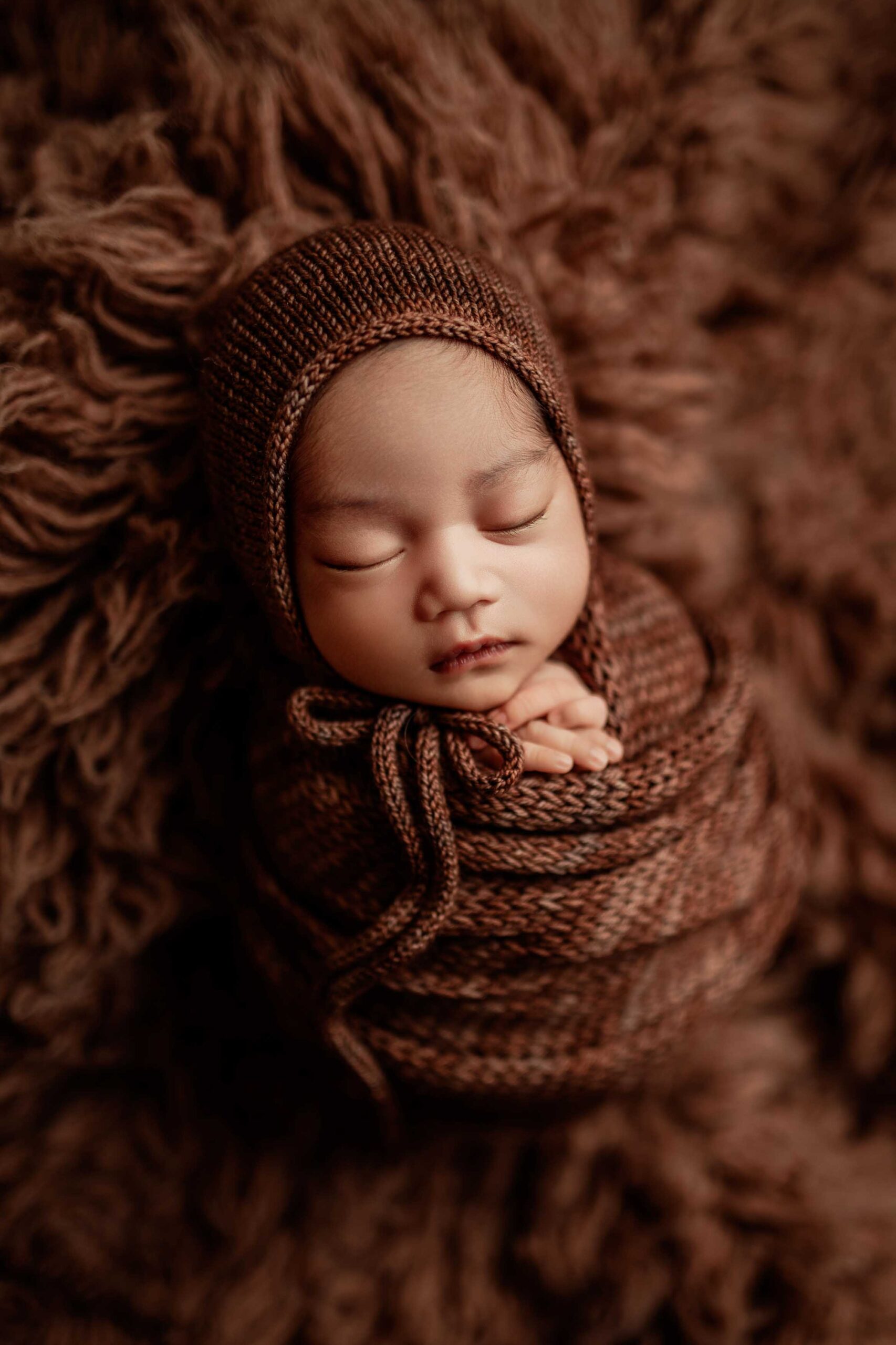 An Adelaide baby wrapped and wearing a bonnet asleep in a brown fur rug.