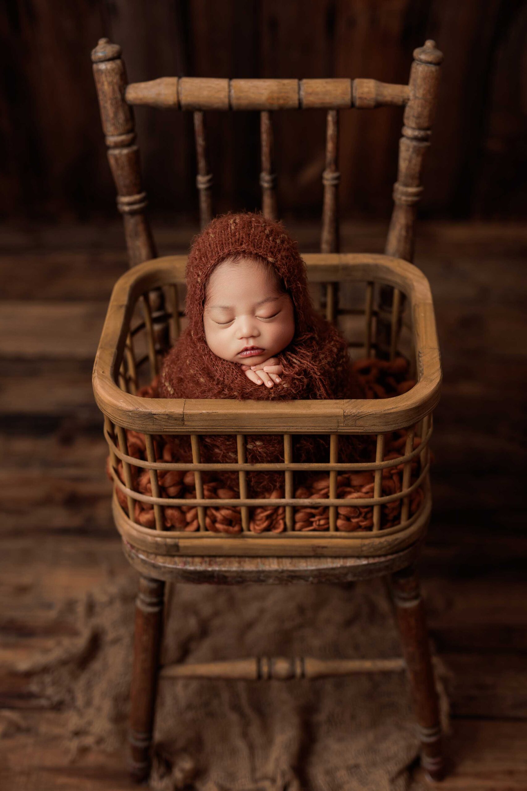An Adelaide newborn baby wrapped and wearing a bonnet asleep and posed upright in a crate on a chair.