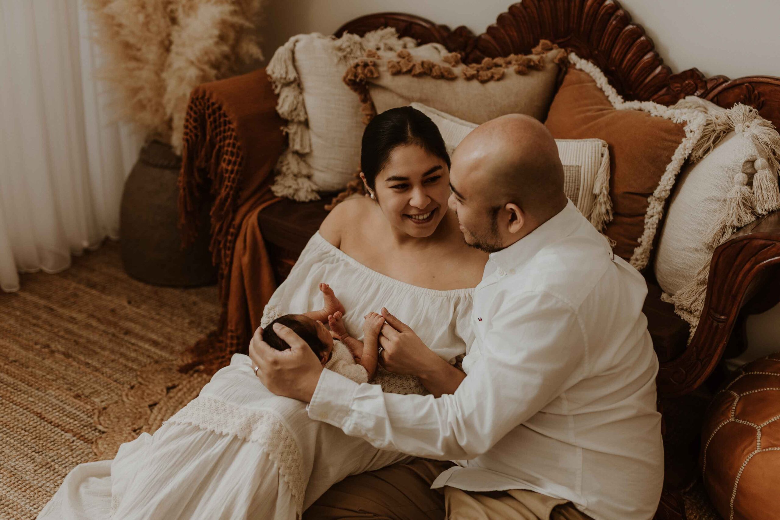 A couple in an Adelaide newborn photography studio sitting on the floor looking at each other while holding their new baby.