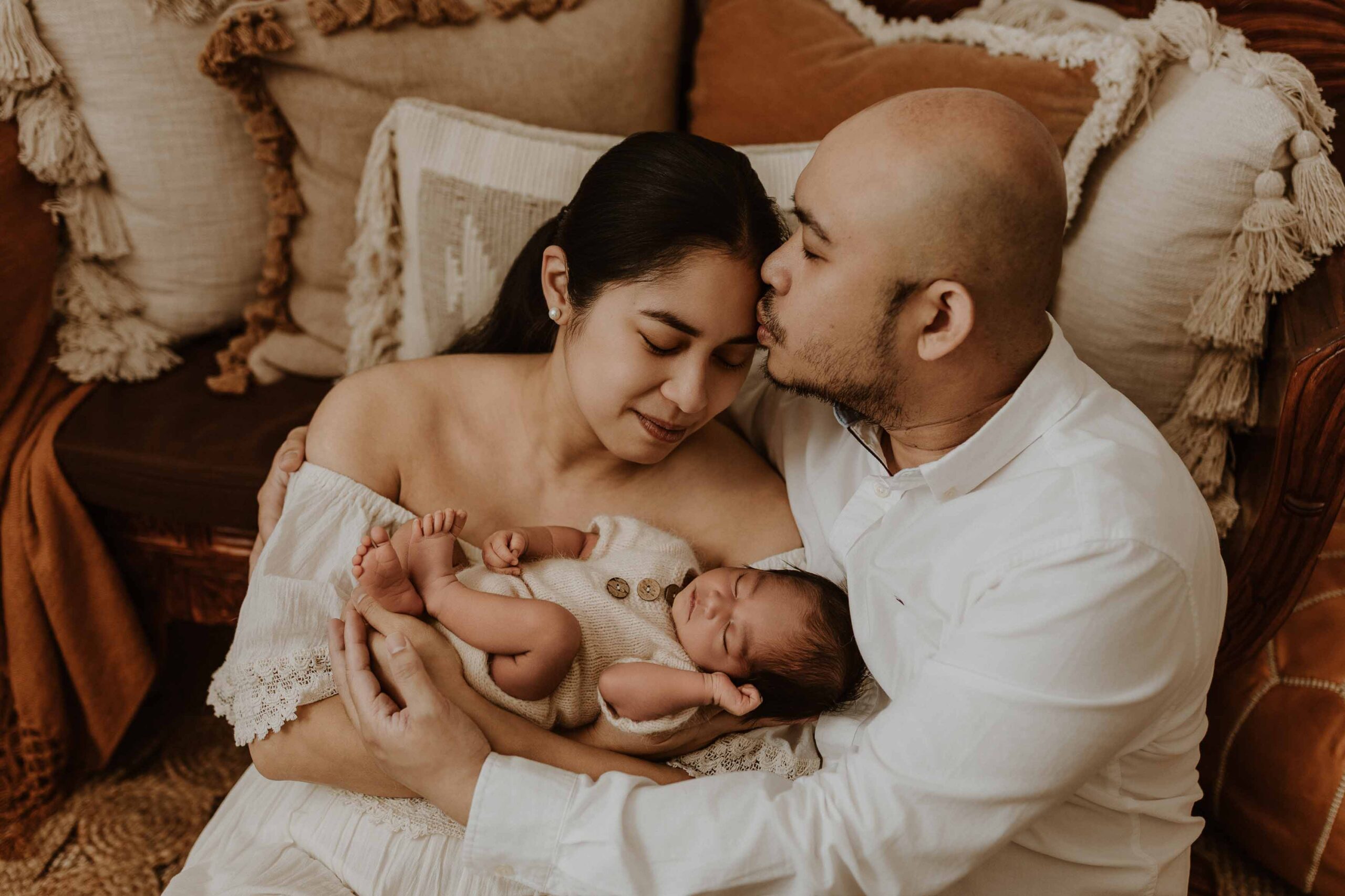 A couple in an Adelaide newborn photography studio sitting on the floor kissing while holding their new baby.
