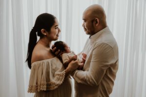 A couple in an Adelaide newborn photography studio smiling at each other while holding their new baby in front of a white curtain.