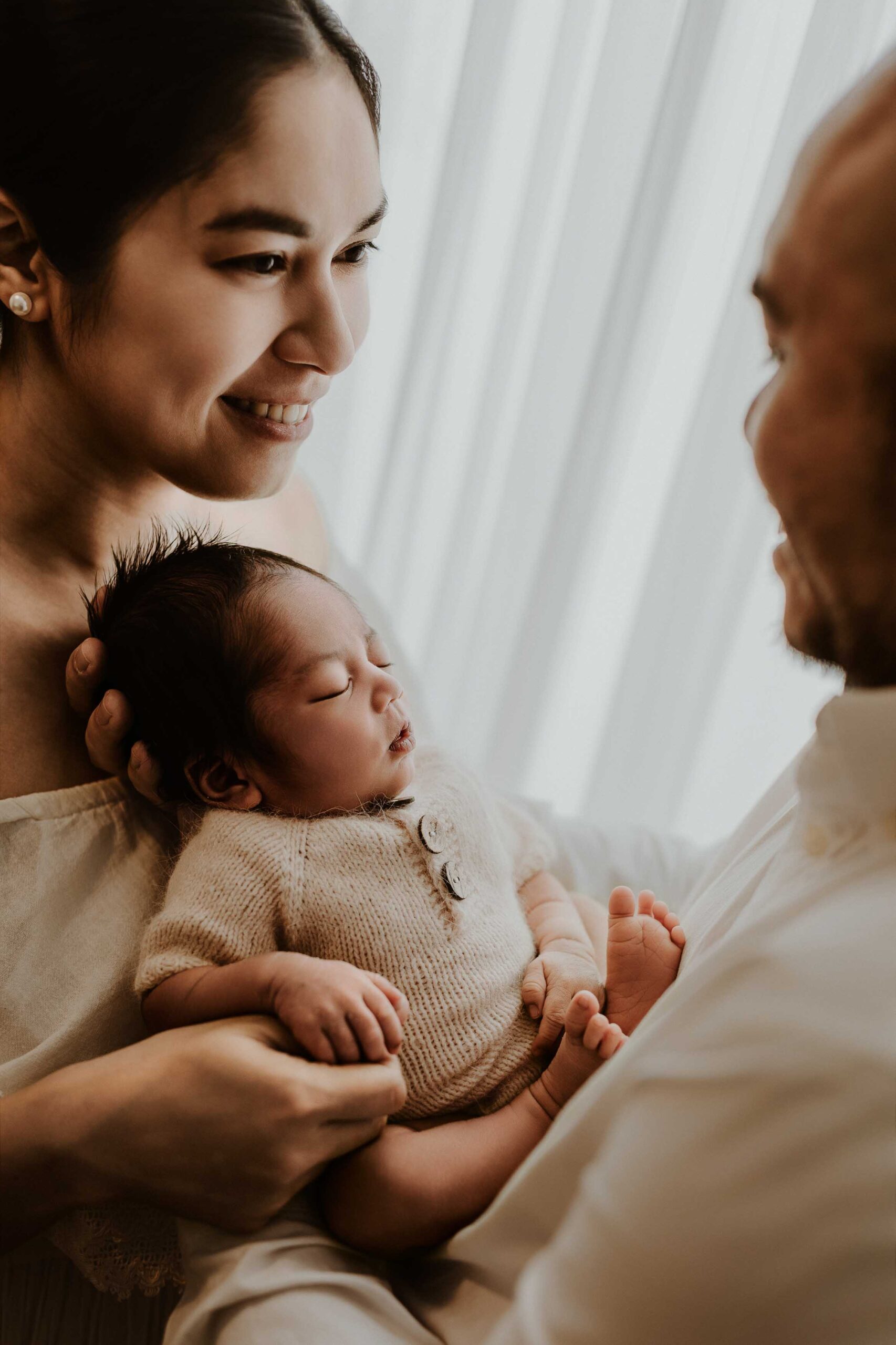 A couple in an Adelaide newborn photography studio smiling while holding their new baby in front of a white curtain.