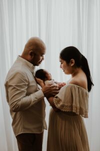 A couple in an Adelaide newborn photography studio smiling at their newborn baby while holding him in front of a white curtain.