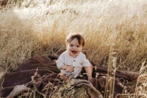 An 8 month old baby boy in a cream shirt sitting on a brown rug in long summer grass.