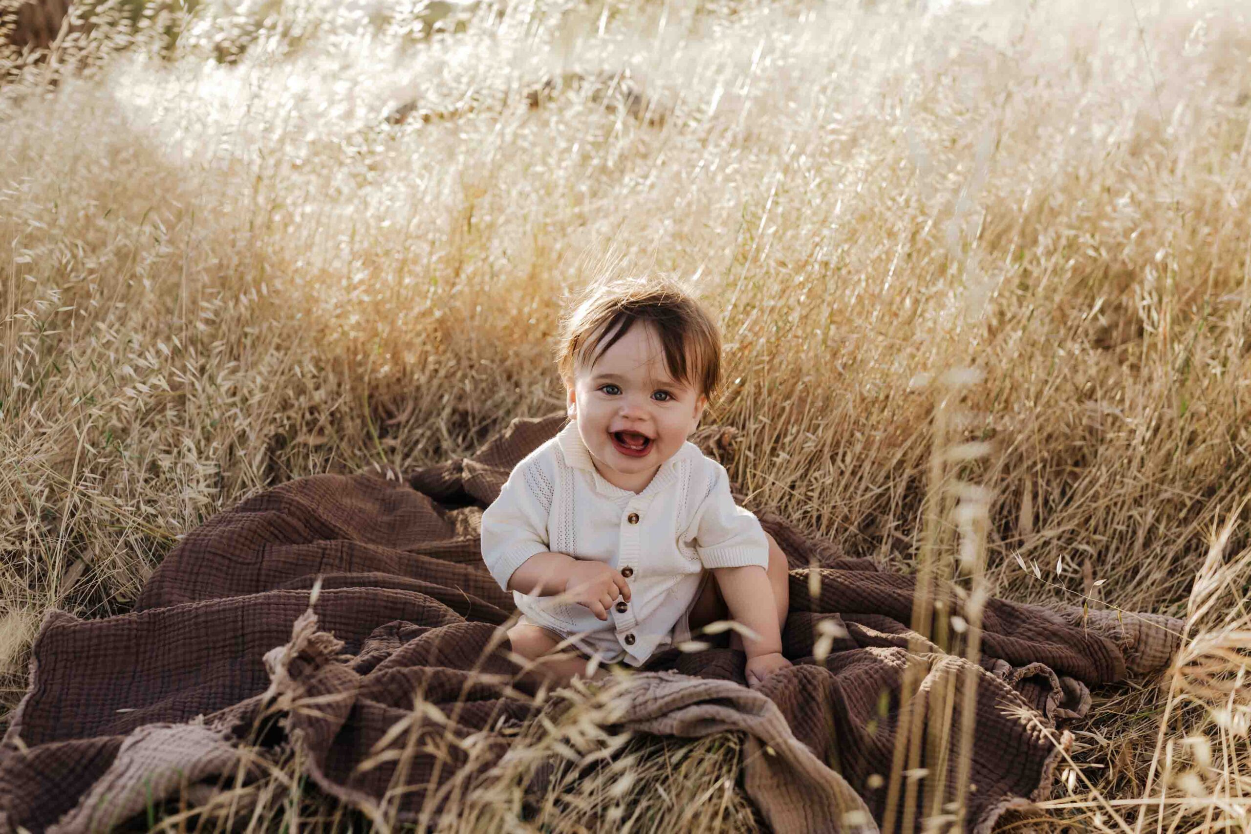 An 8 month old baby boy in a cream shirt sitting on a brown rug in long summer grass.
