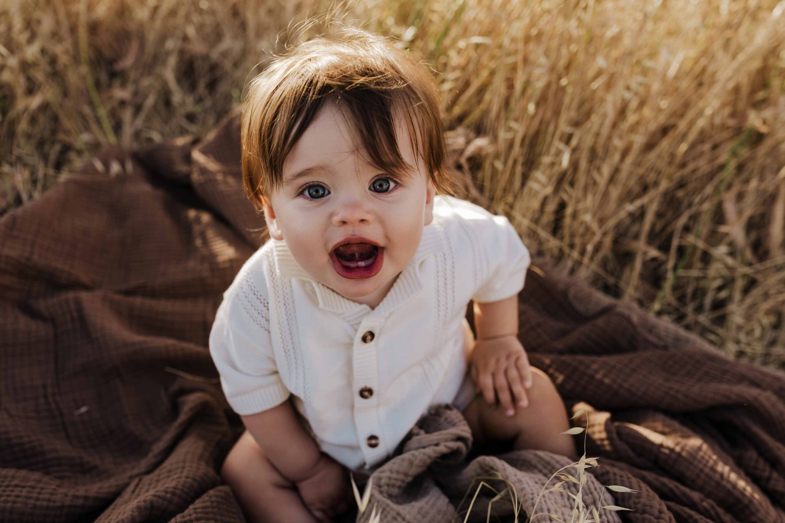 An 8 month old baby boy in a cream shirt sitting on a brown rug in long summer grass.
