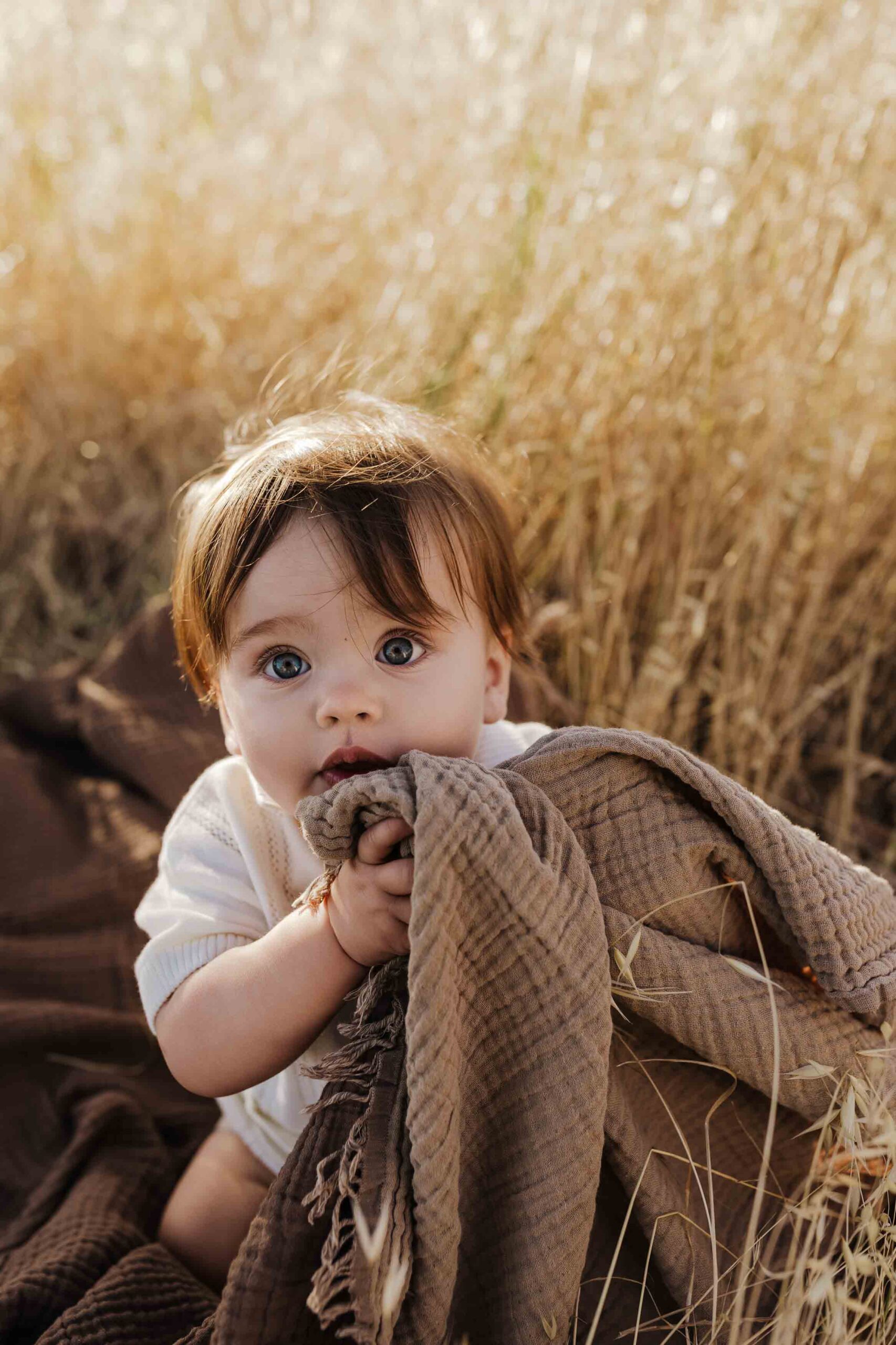 An 8 month old baby boy in a cream shirt sitting on a brown rug in long summer grass.
