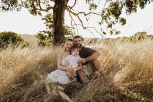 An Adelaide mother and father sitting with their 8 month old son outside in a field in front of a tree.