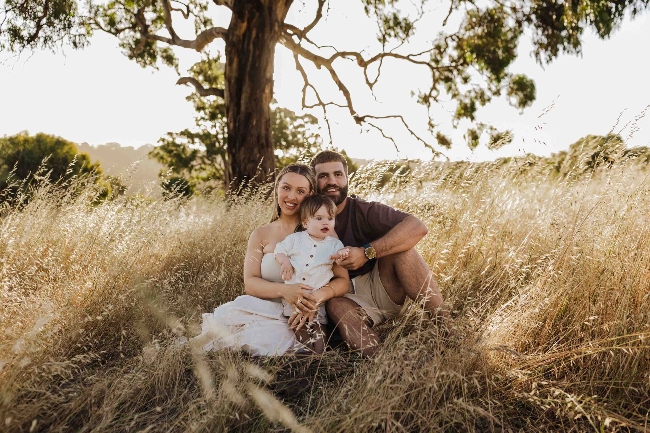 An Adelaide mother and father sitting with their 8 month old son outside in a field in front of a tree.