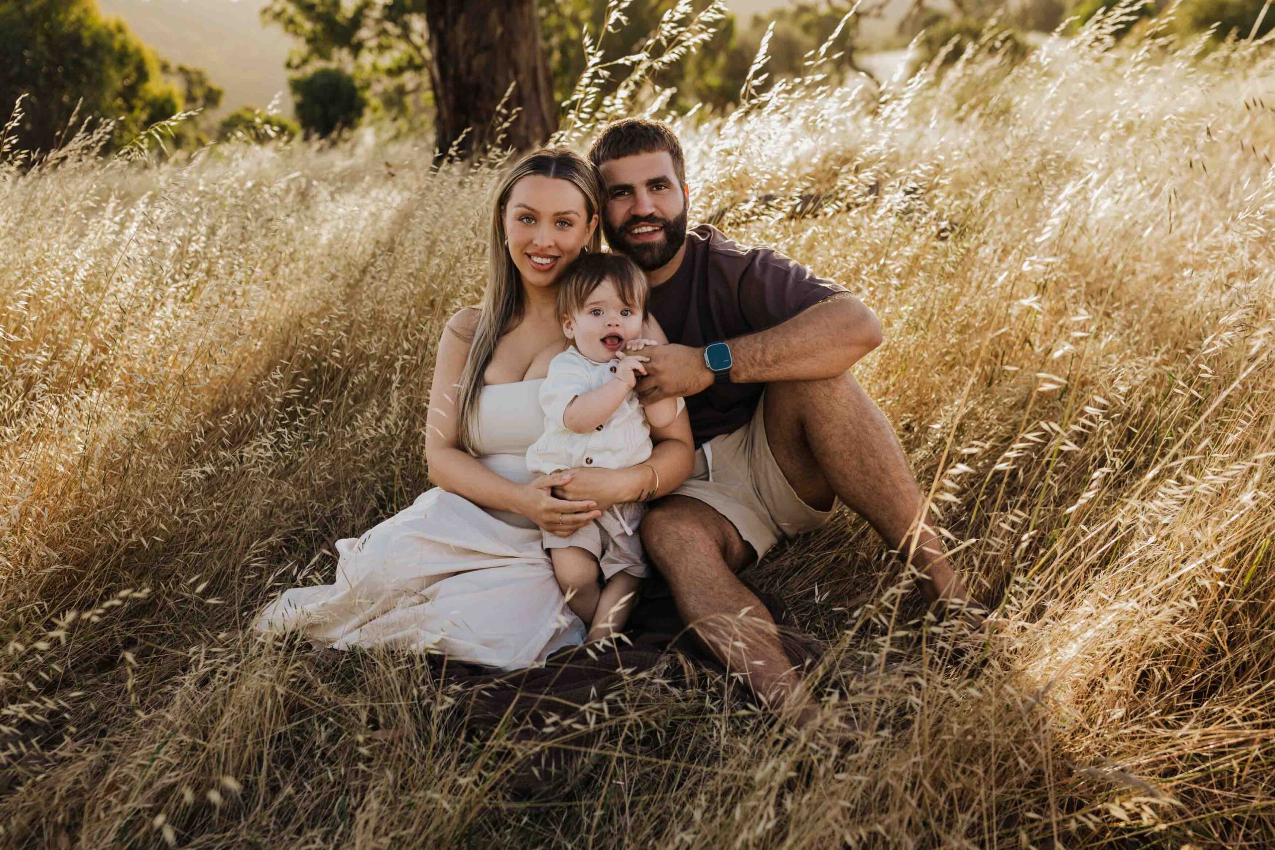 An Adelaide mother and father sitting with their 8 month old son outside in a field in front of a tree.
