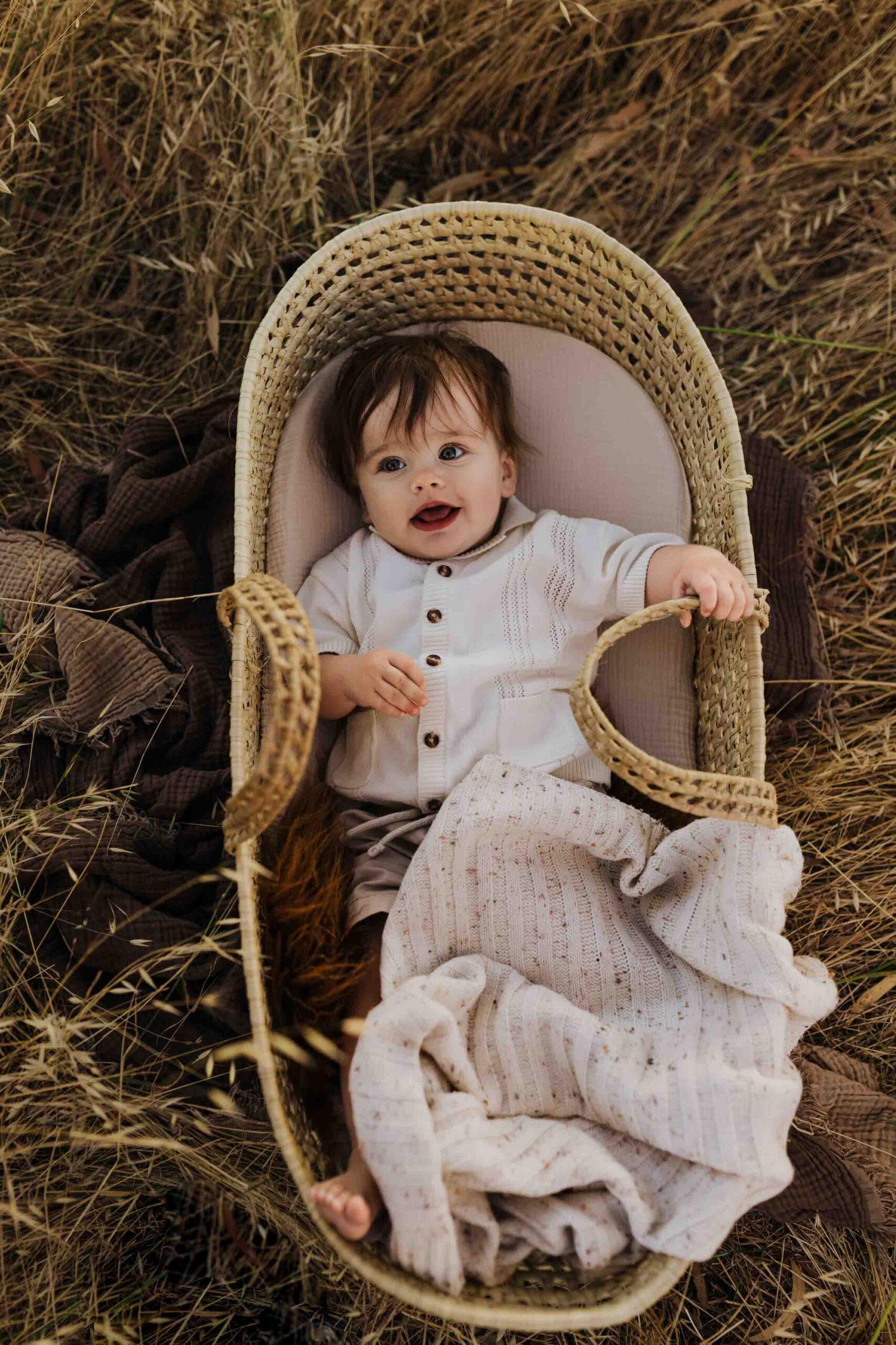 An 8 month old baby boy in a cream shirt laying in a woven mosses basket in long summer grass.