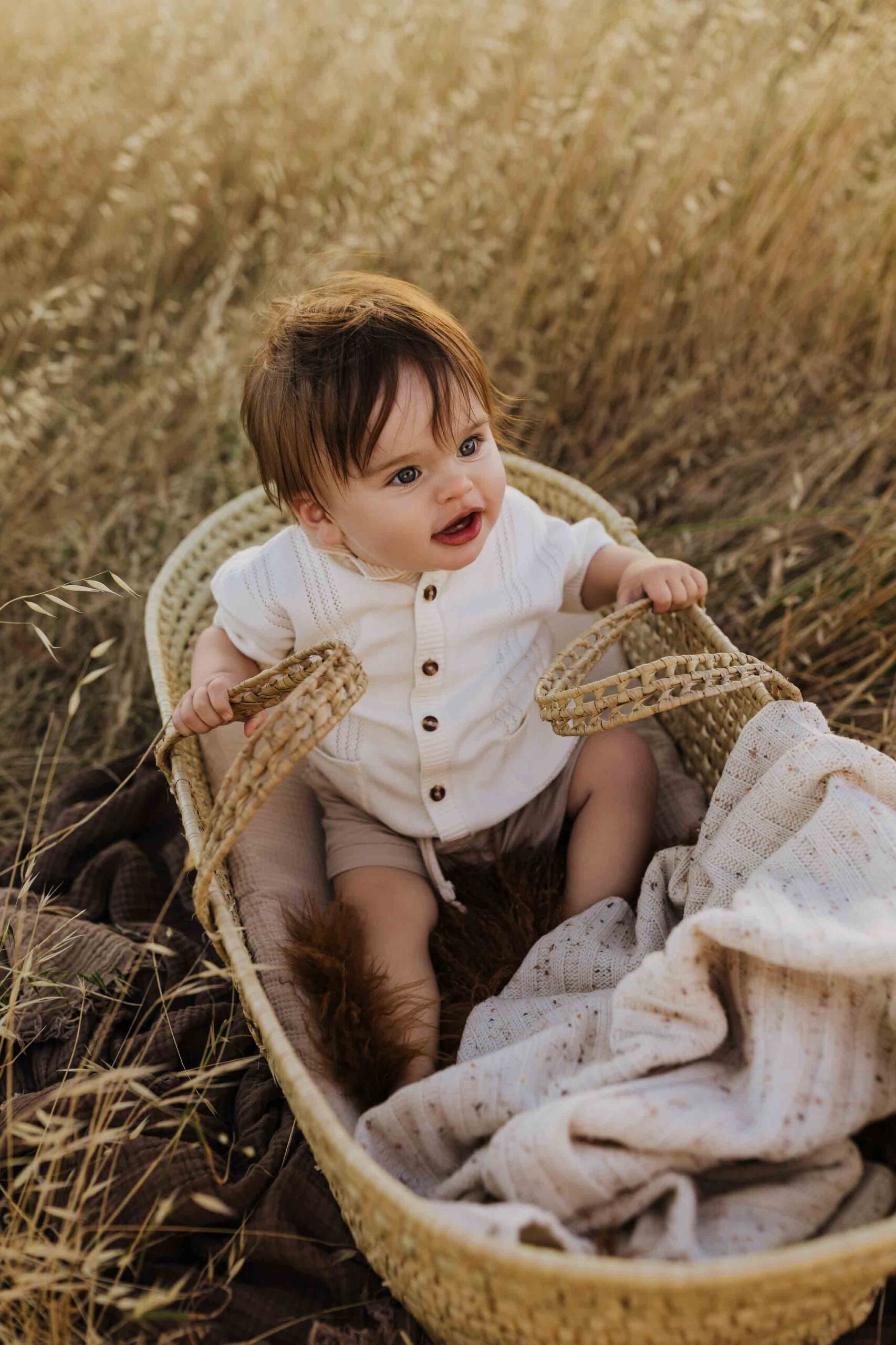 An 8 month old baby boy in a cream shirt sitting in a woven mosses basket in long summer grass.