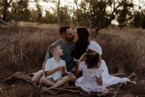 A family of 4 sitting in the grass with mum and dad kissing..
