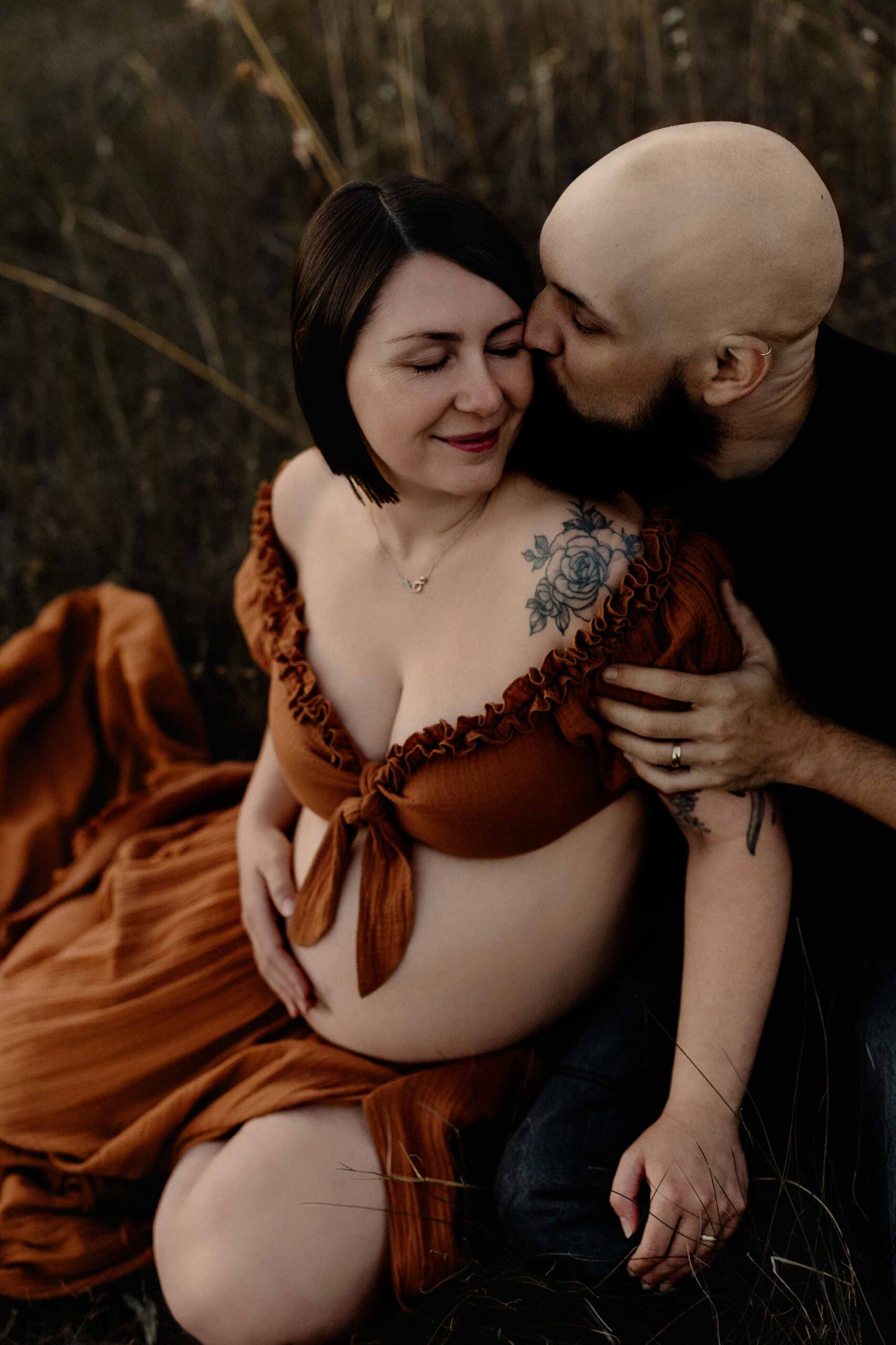A pregnant mother and her partner who is kissing her cheek sitting in grass.