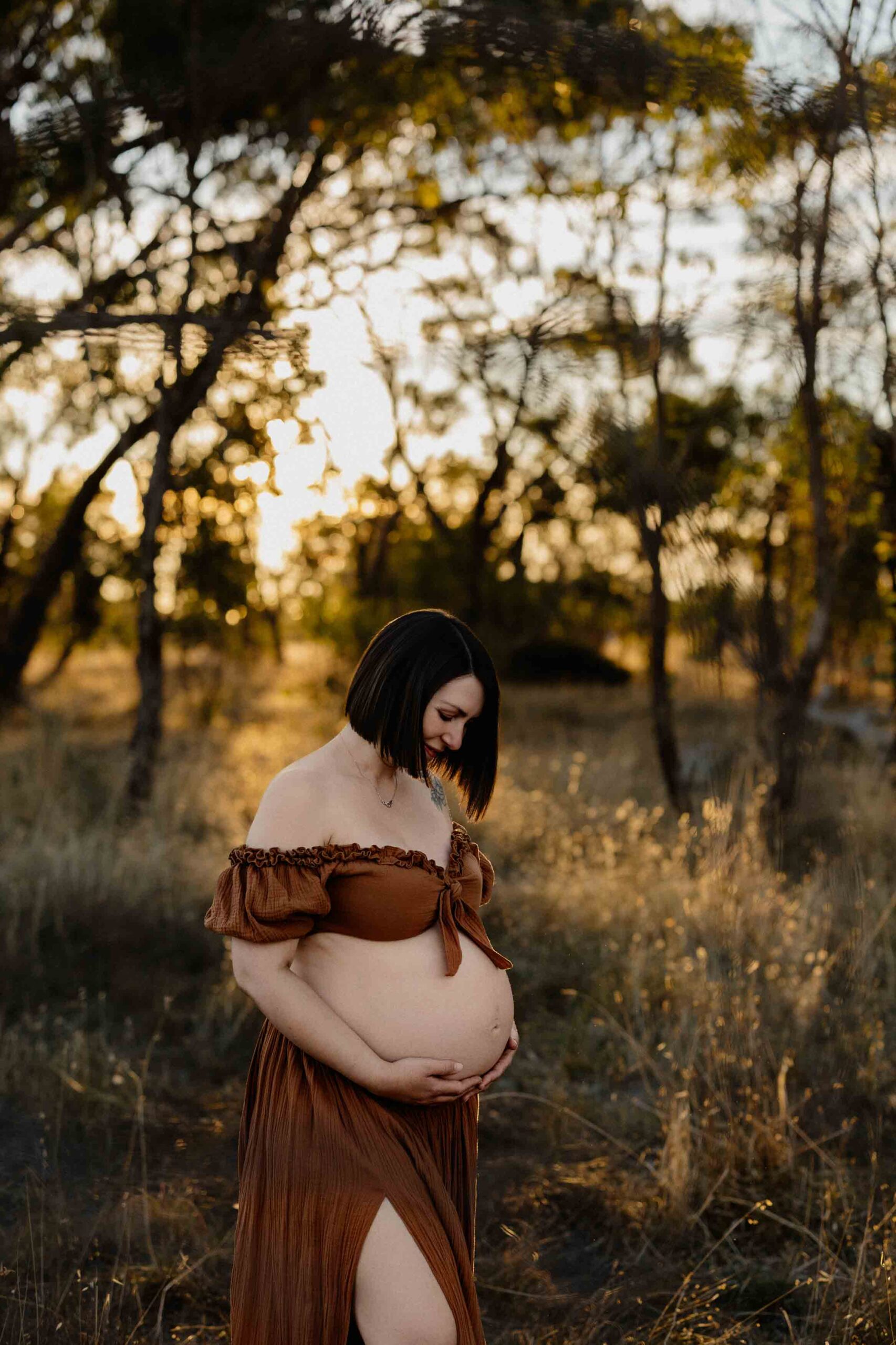 A mum from Adelaide looking at her pregnant belly wearing a 2 piece rust outfit at sunset.