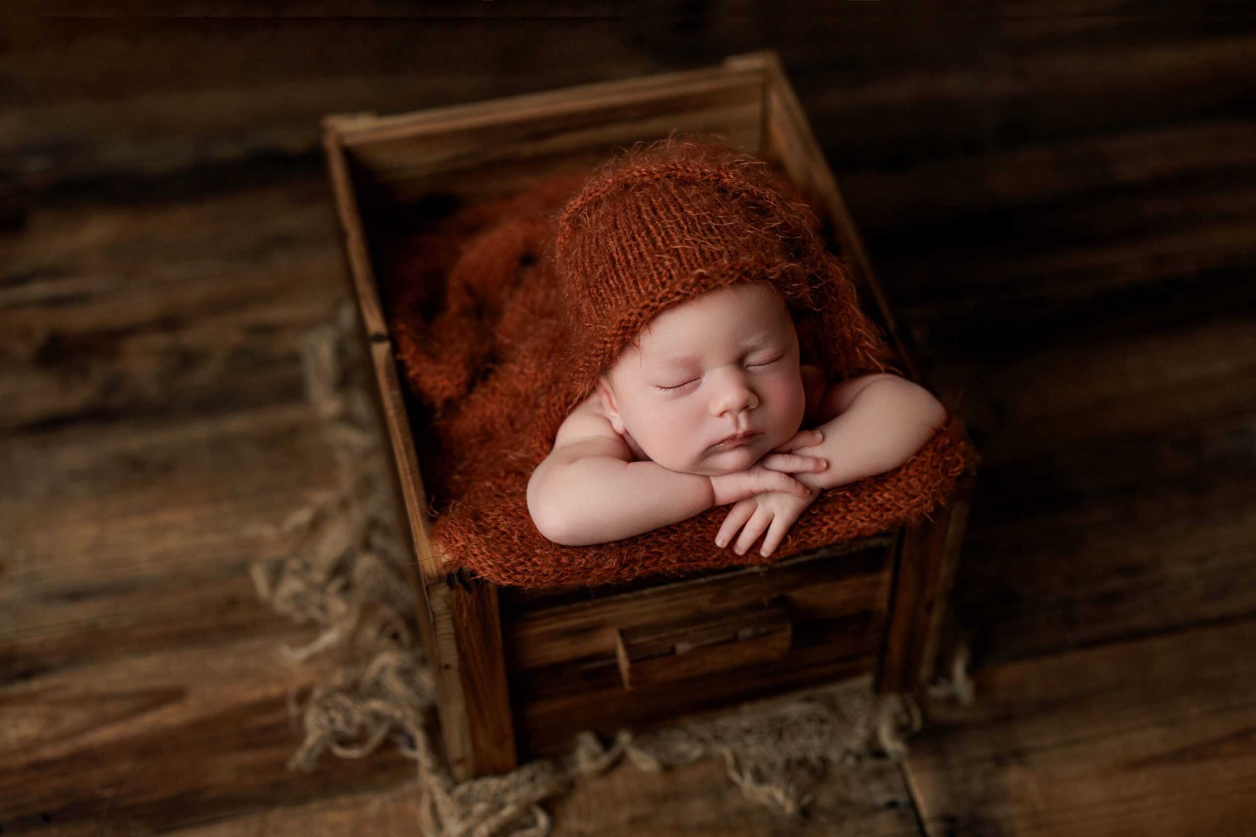 A sleeping baby wearing a rust-coloured knitted bonnet rests with folded arms on the edge of a wooden box, surrounded by matching fabric, on a rustic wooden floor—capturing the charm of Adelaide baby photos.