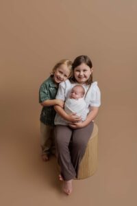Three children pose together on a neutral brown background; an older girl sits on a round stool holding a swaddled baby, whilst a smiling boy stands beside her with his arm around her shoulders in this charming Adelaide baby photoshoot.