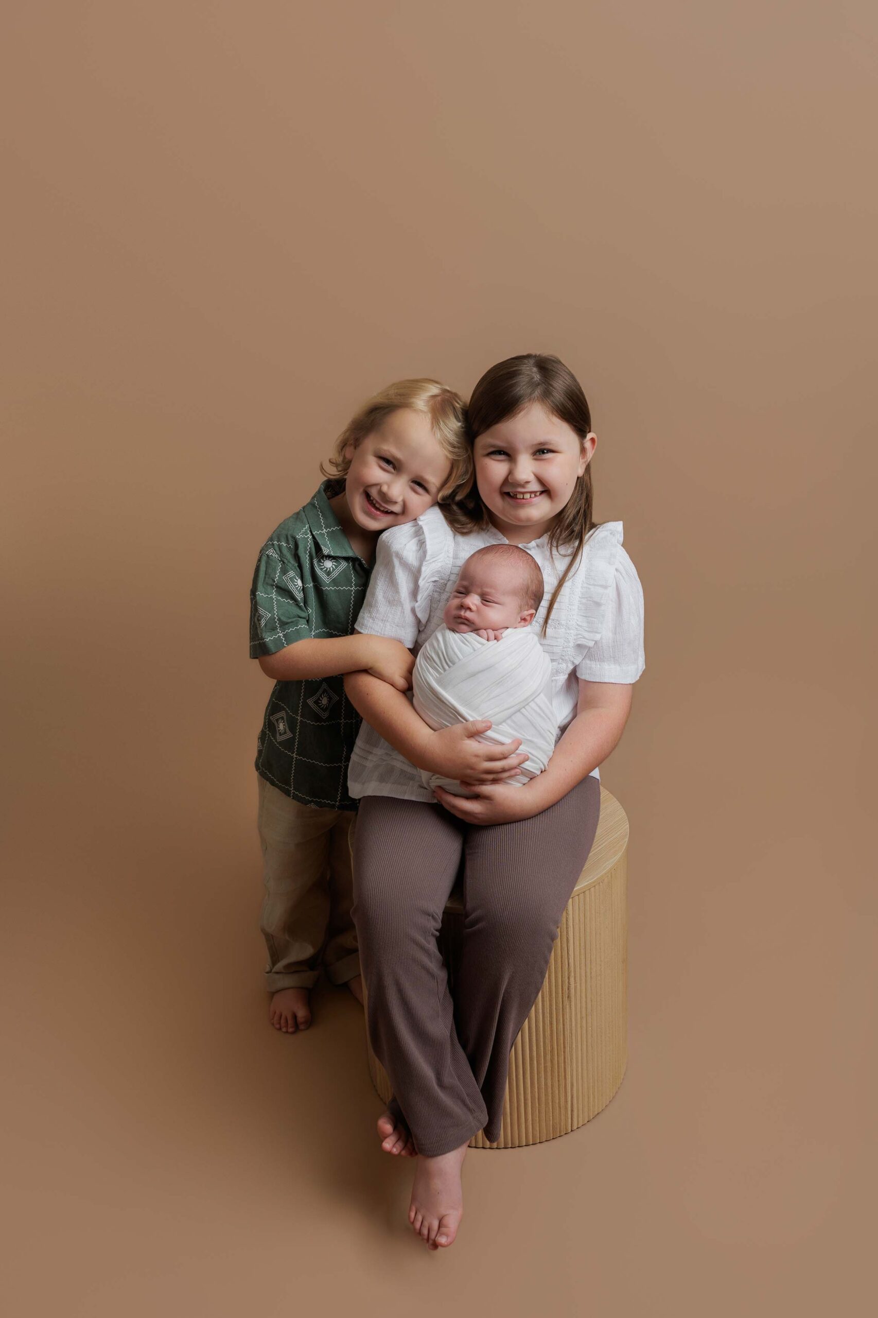 Three children pose together on a neutral brown background; an older girl sits on a round stool holding a swaddled baby, whilst a smiling boy stands beside her with his arm around her shoulders in this charming Adelaide baby photoshoot.
