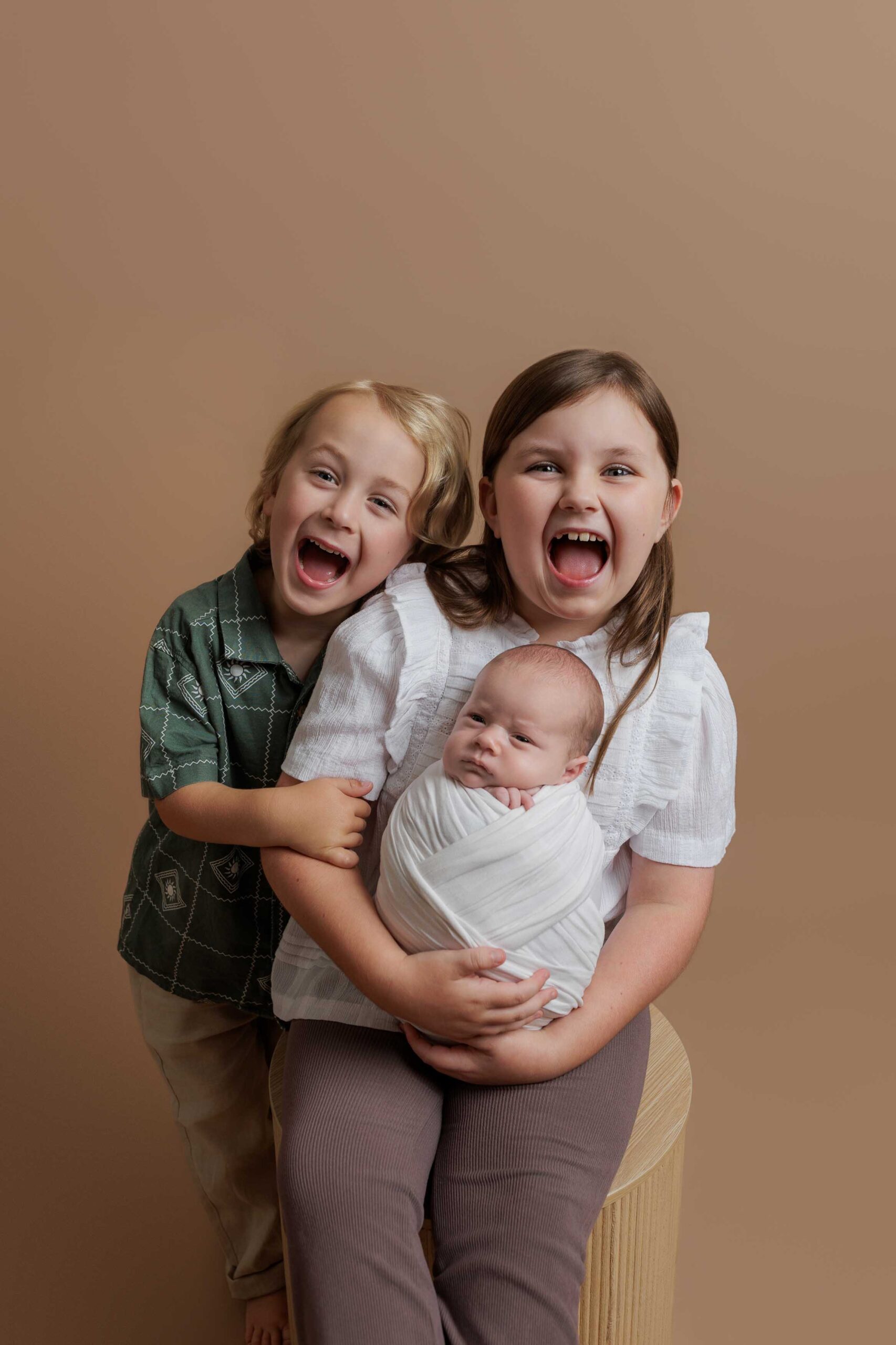 Two smiling children, a boy and a girl, pose happily against a brown background. The girl sits on a stool holding a swaddled baby in her lap, whilst the boy hugs them both—capturing the warmth of Adelaide baby photos.