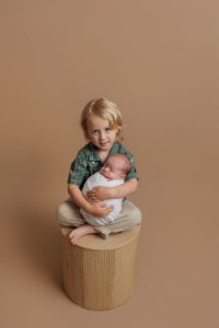 A young child with fair hair sits barefoot on a round stool, gently holding a swaddled newborn baby in their lap. This sweet moment, set against a plain beige background, captures the warmth of Adelaide baby photos.