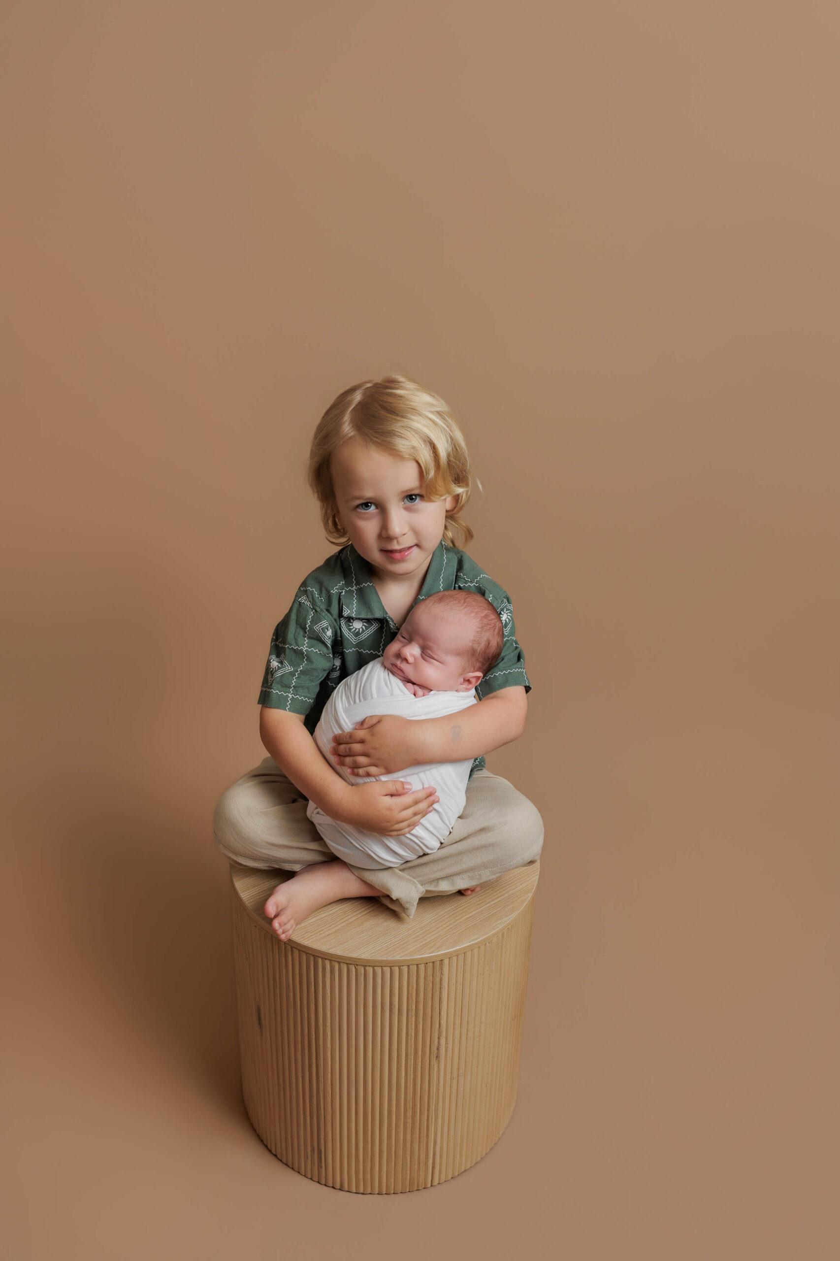 A young child with fair hair sits barefoot on a round stool, gently holding a swaddled newborn baby in their lap. This sweet moment, set against a plain beige background, captures the warmth of Adelaide baby photos.