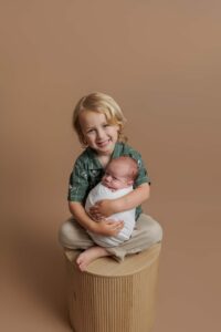 A young child with blonde hair, dressed in a green shirt and beige trousers, sits on a round stool and smiles whilst gently holding a swaddled baby in their lap. Perfect for Adelaide baby photos, the background is a solid light brown colour.