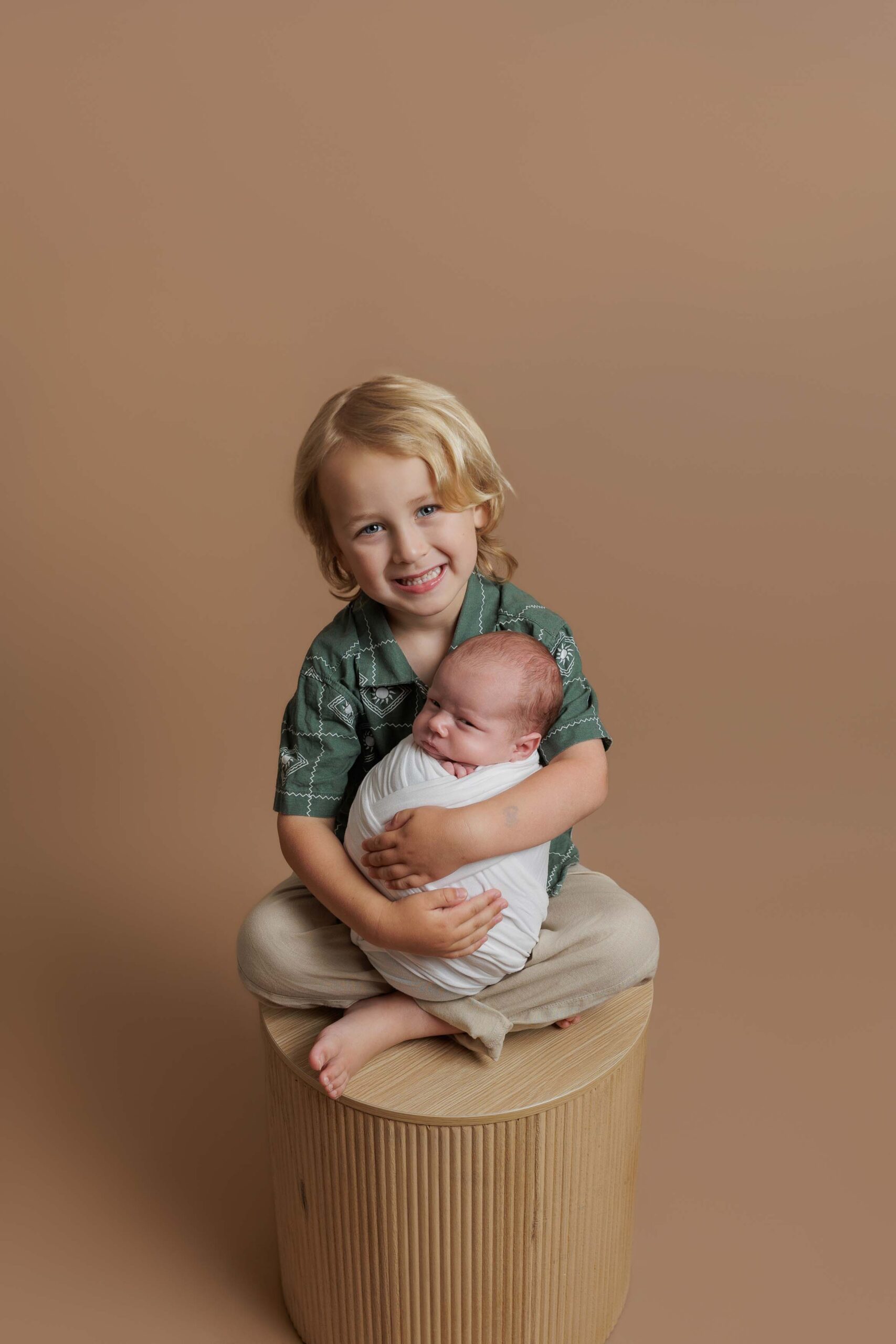 A young child with blonde hair, dressed in a green shirt and beige trousers, sits on a round stool and smiles whilst gently holding a swaddled baby in their lap. Perfect for Adelaide baby photos, the background is a solid light brown colour.