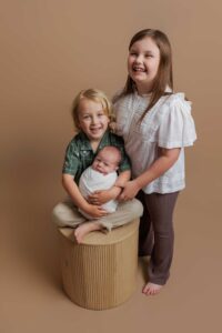 Three smiling children pose against a beige background for sweet Adelaide baby photos. An older girl stands beside a young boy sitting on a wooden cylinder, holding a swaddled baby in his lap. All three look happy and close together.