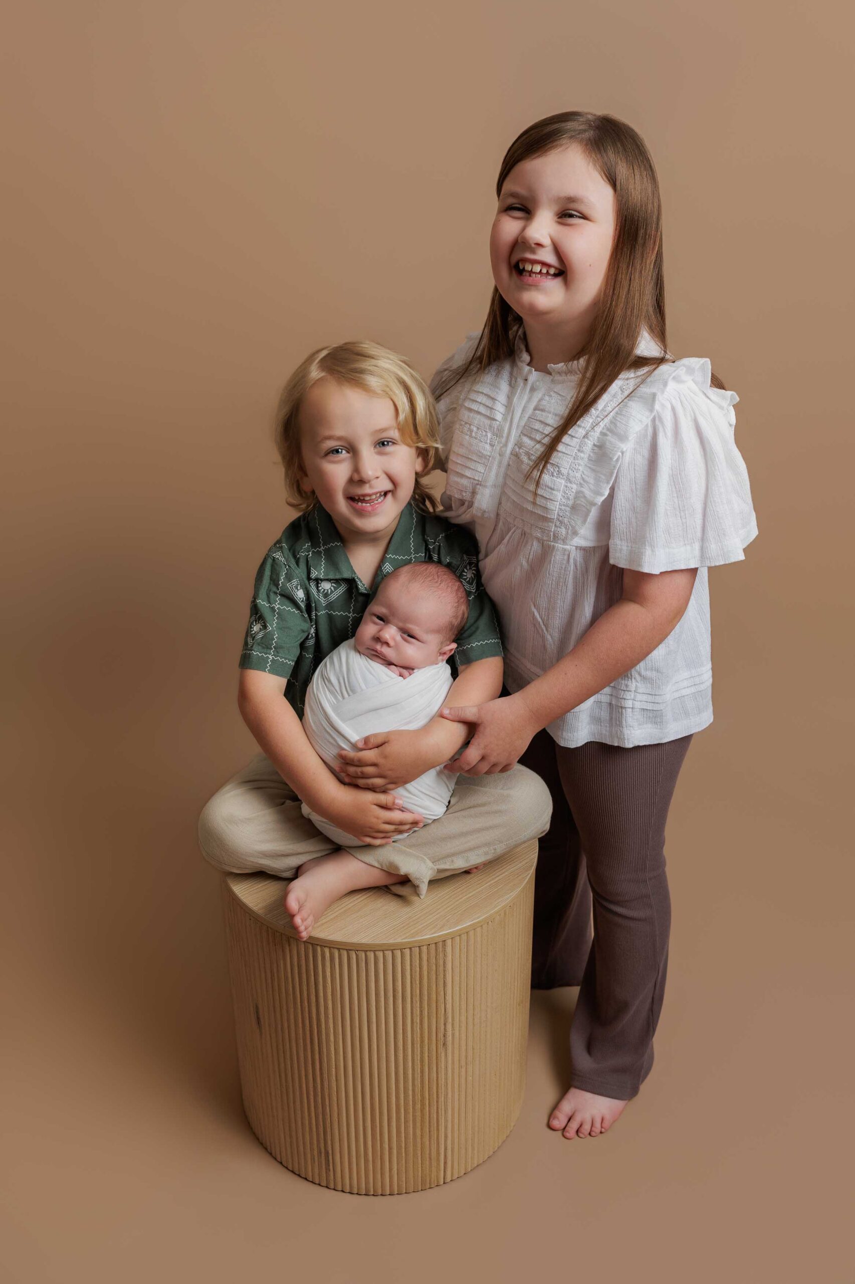 Three smiling children pose against a beige background for sweet Adelaide baby photos. An older girl stands beside a young boy sitting on a wooden cylinder, holding a swaddled baby in his lap. All three look happy and close together.