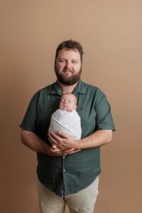 A bearded man in a green shirt holds a newborn wrapped in a white blanket, standing against a plain beige background. Their calm, content expressions make this a timeless example of Adelaide baby photos.