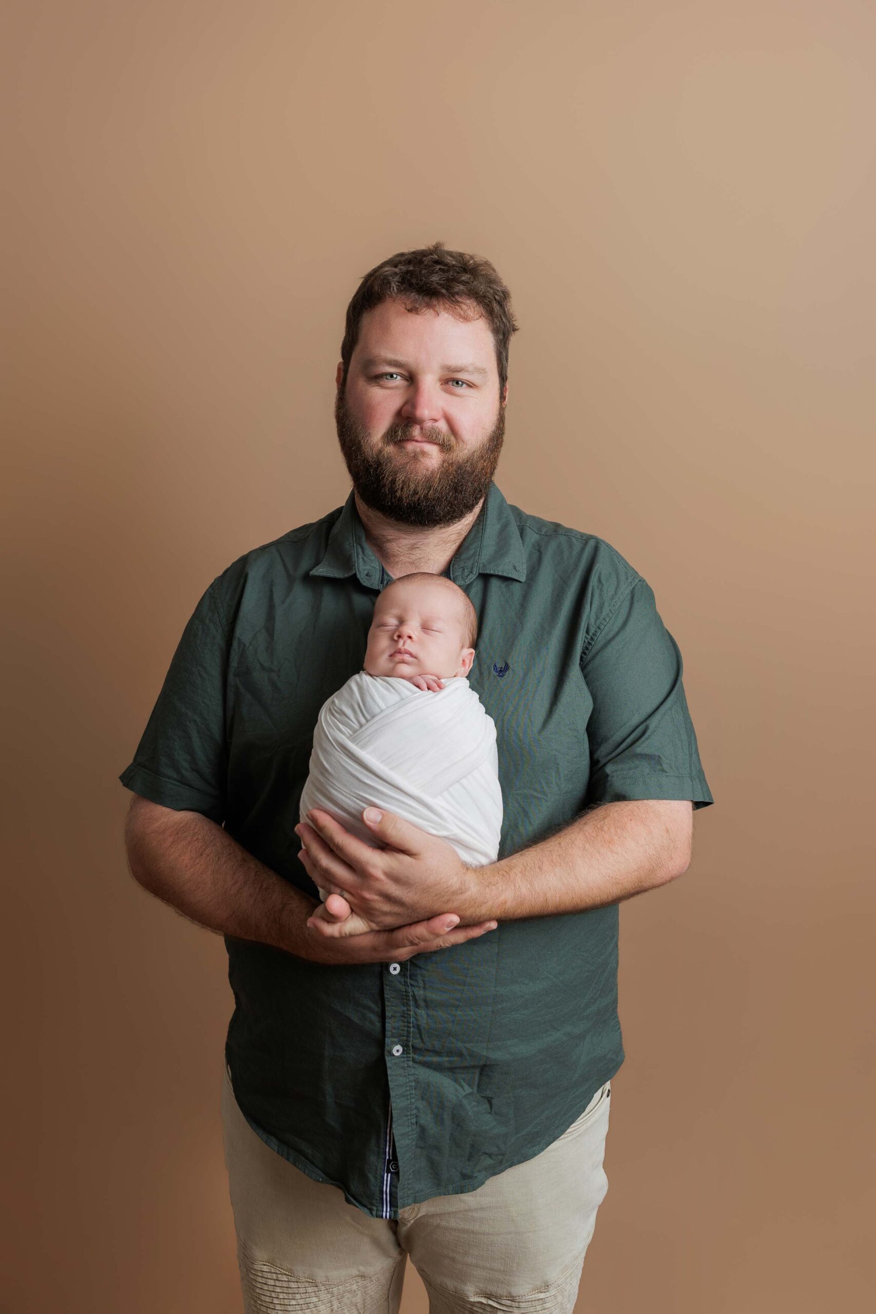 A bearded man in a green shirt holds a newborn wrapped in a white blanket, standing against a plain beige background. Their calm, content expressions make this a timeless example of Adelaide baby photos.