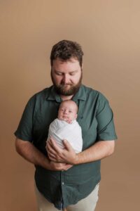 A bearded man in a green shirt gently cradles a swaddled baby in his arms, standing against a plain beige background—an intimate moment captured in classic Adelaide baby photos style.