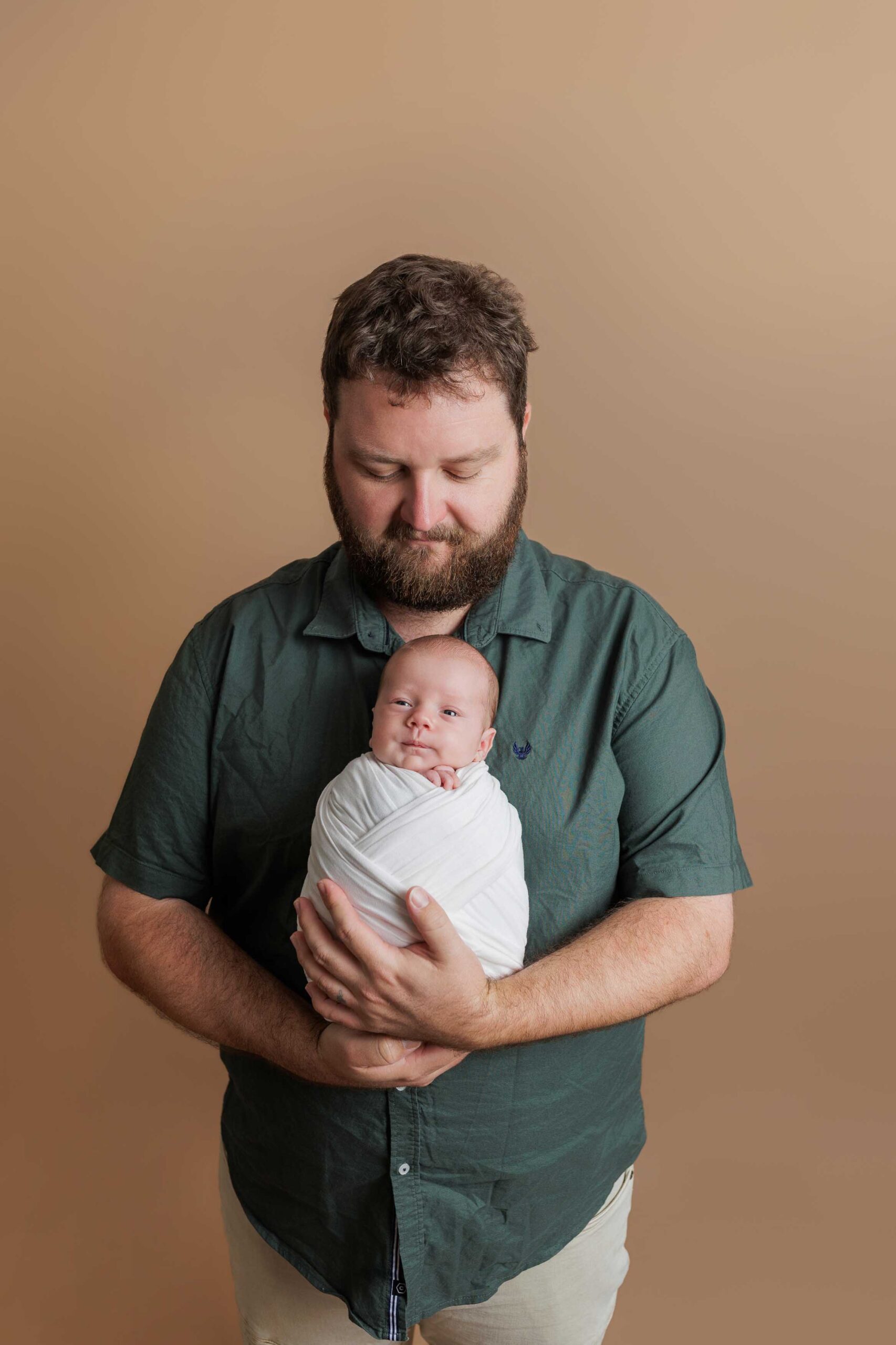 A bearded man in a green shirt gently cradles a swaddled baby in his arms, standing against a plain beige background—an intimate moment captured in classic Adelaide baby photos style.
