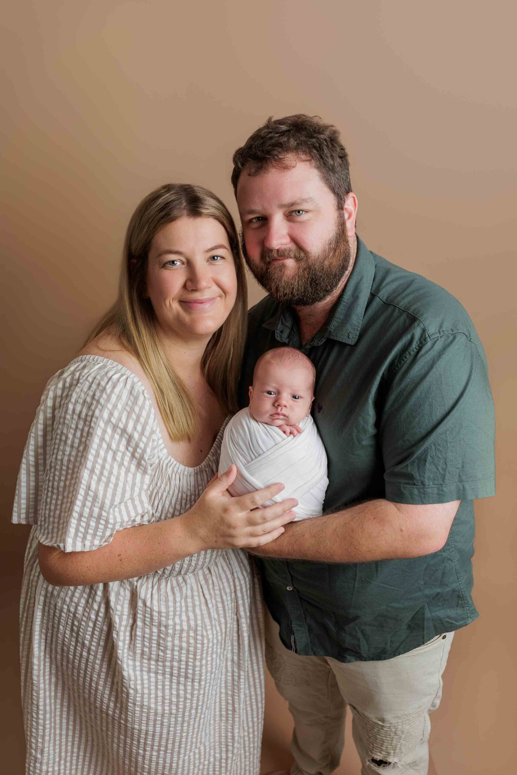A woman and man stand close together, smiling, as they hold a newborn baby swaddled in a white blanket. Capturing Adelaide baby photos, the scene features a soft light brown background perfect for this precious family moment.
