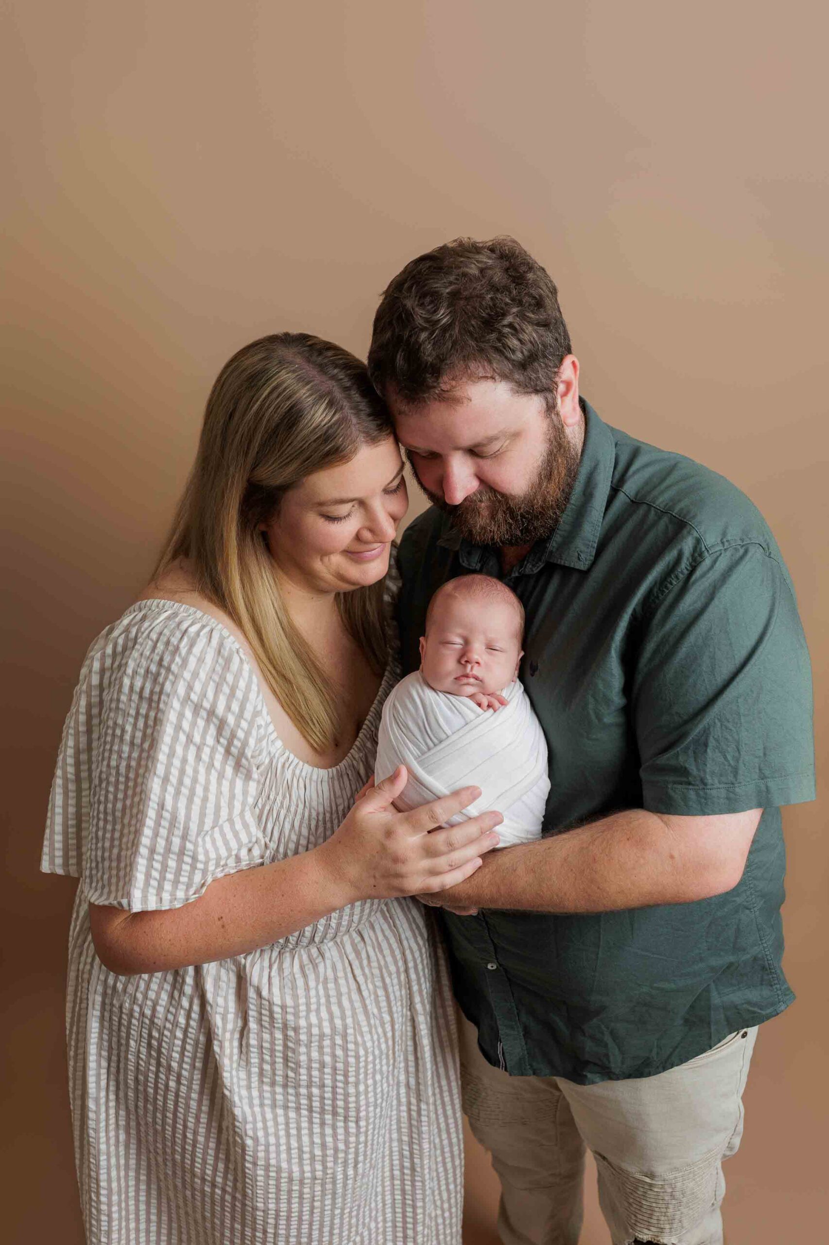 A couple stands closely together, lovingly holding a swaddled newborn baby. The woman wears a light striped dress and the man a dark green shirt. Capturing peaceful Adelaide baby photos, all three pose against a soft beige background, appearing contented.