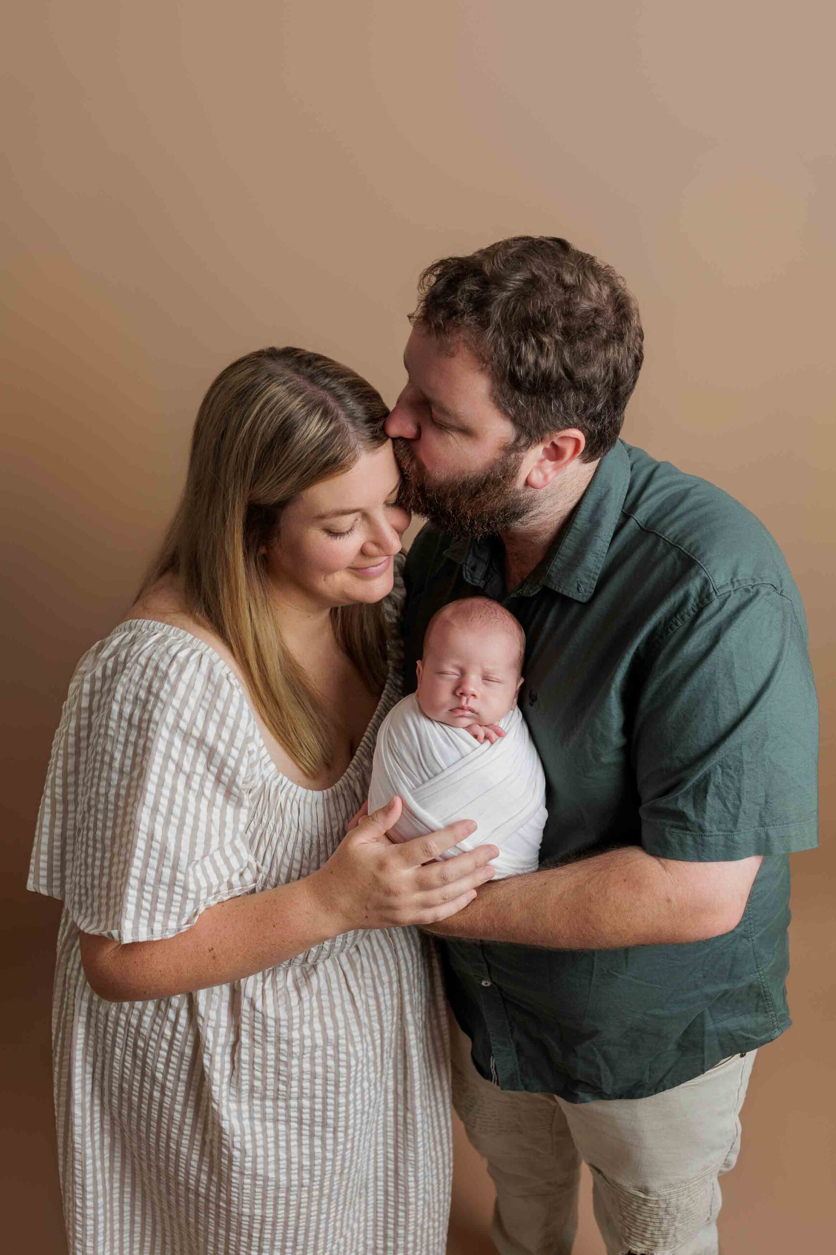 A mother and father lovingly hold their newborn swaddled in a white blanket. The father kisses the mother's head while she smiles, all standing close together against a plain beige background, capturing tender Adelaide baby photographs.