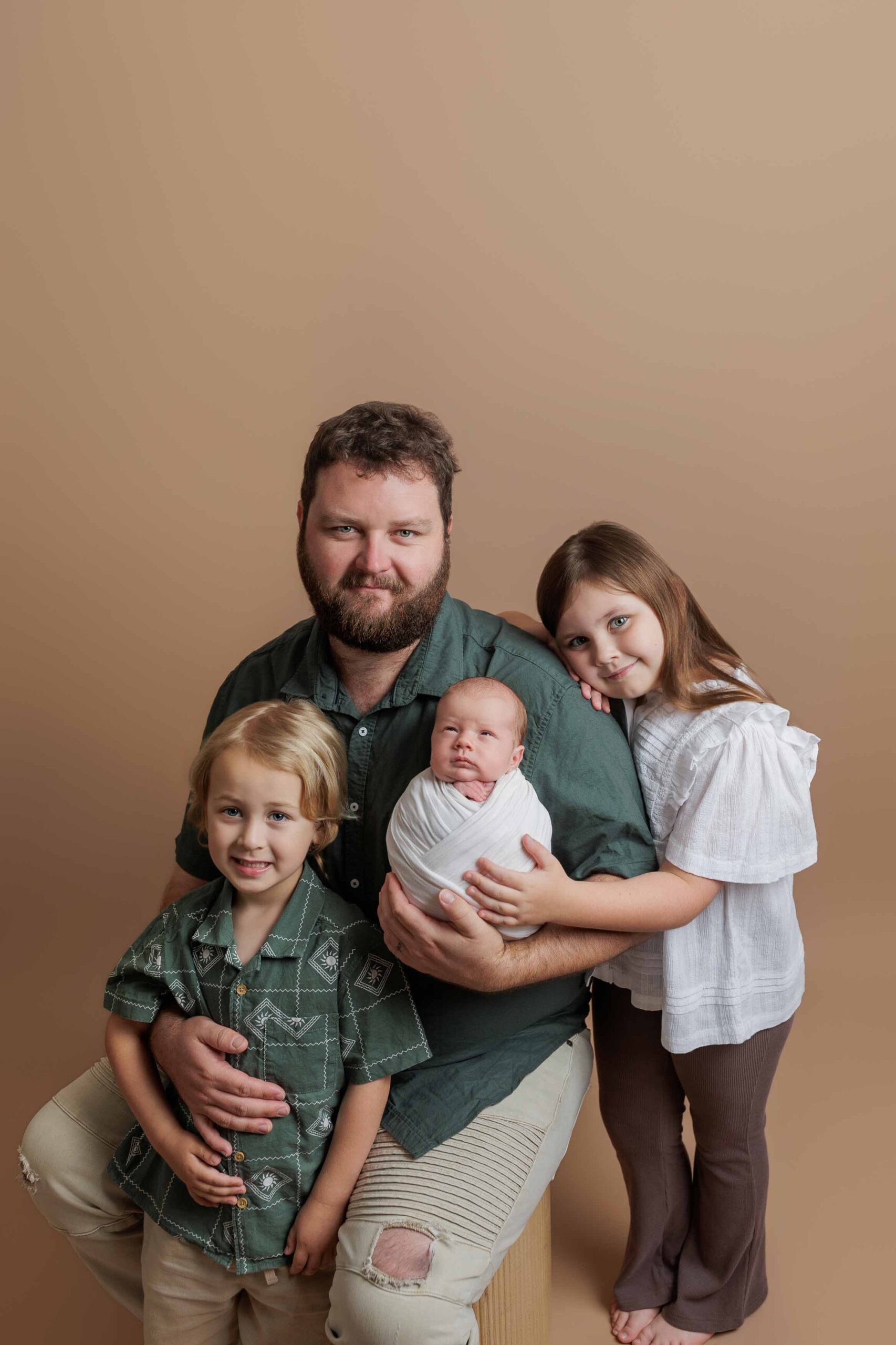 A bearded man sits holding a newborn wrapped in white, with a young boy beside him and a young girl leaning on his shoulder, all smiling. This heartwarming Adelaide baby photo captures family joy against a soft beige background.