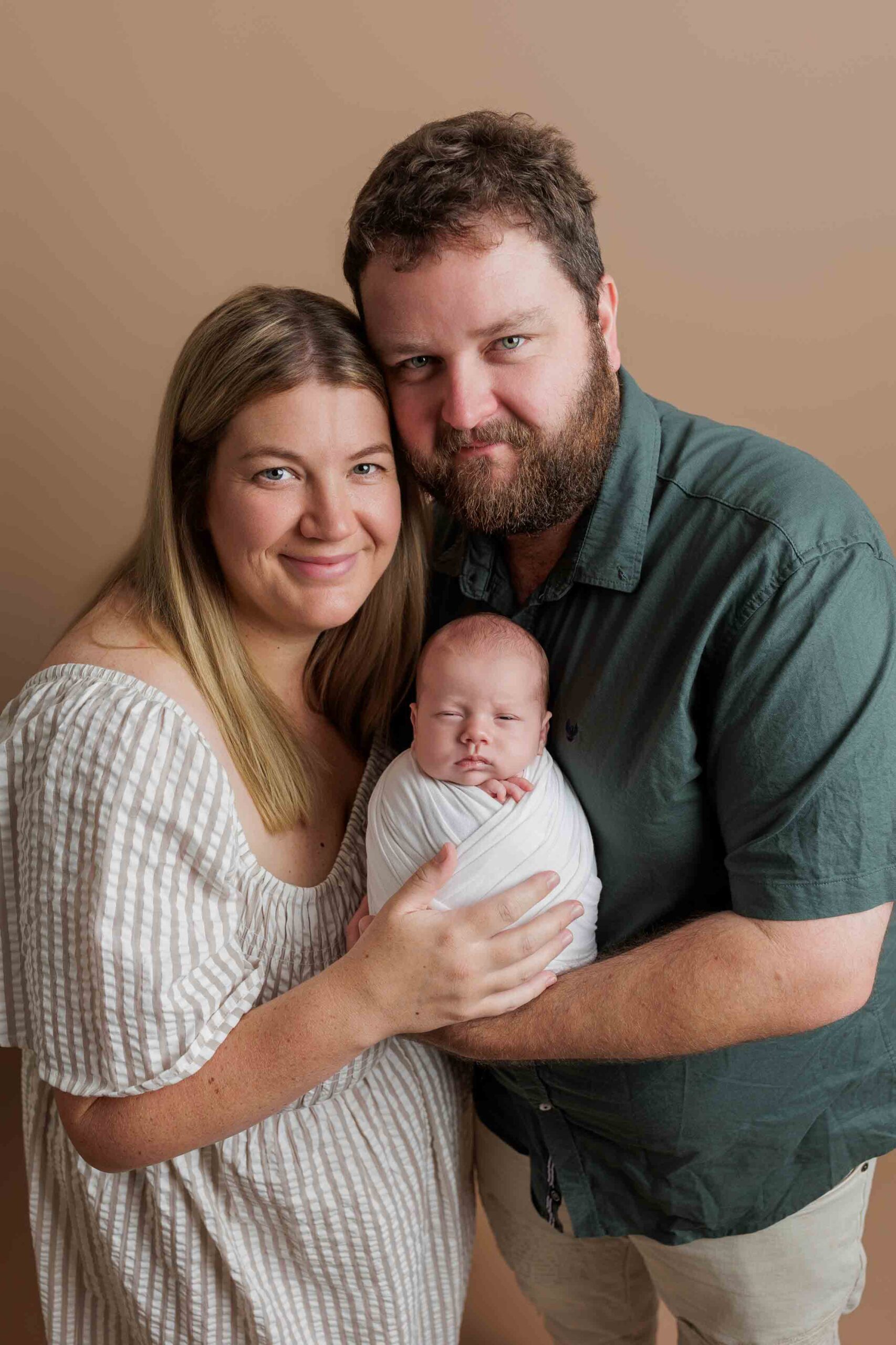 A smiling woman and man hold a newborn baby wrapped in a white blanket. The family stands closely together in front of a plain beige background, capturing the warmth of Adelaide baby photos.