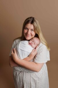 A woman with long blonde hair smiles gently whilst holding a sleeping newborn wrapped in a white blanket against a light brown background. She is wearing a light-coloured, striped dress—perfect for timeless Adelaide baby photos.