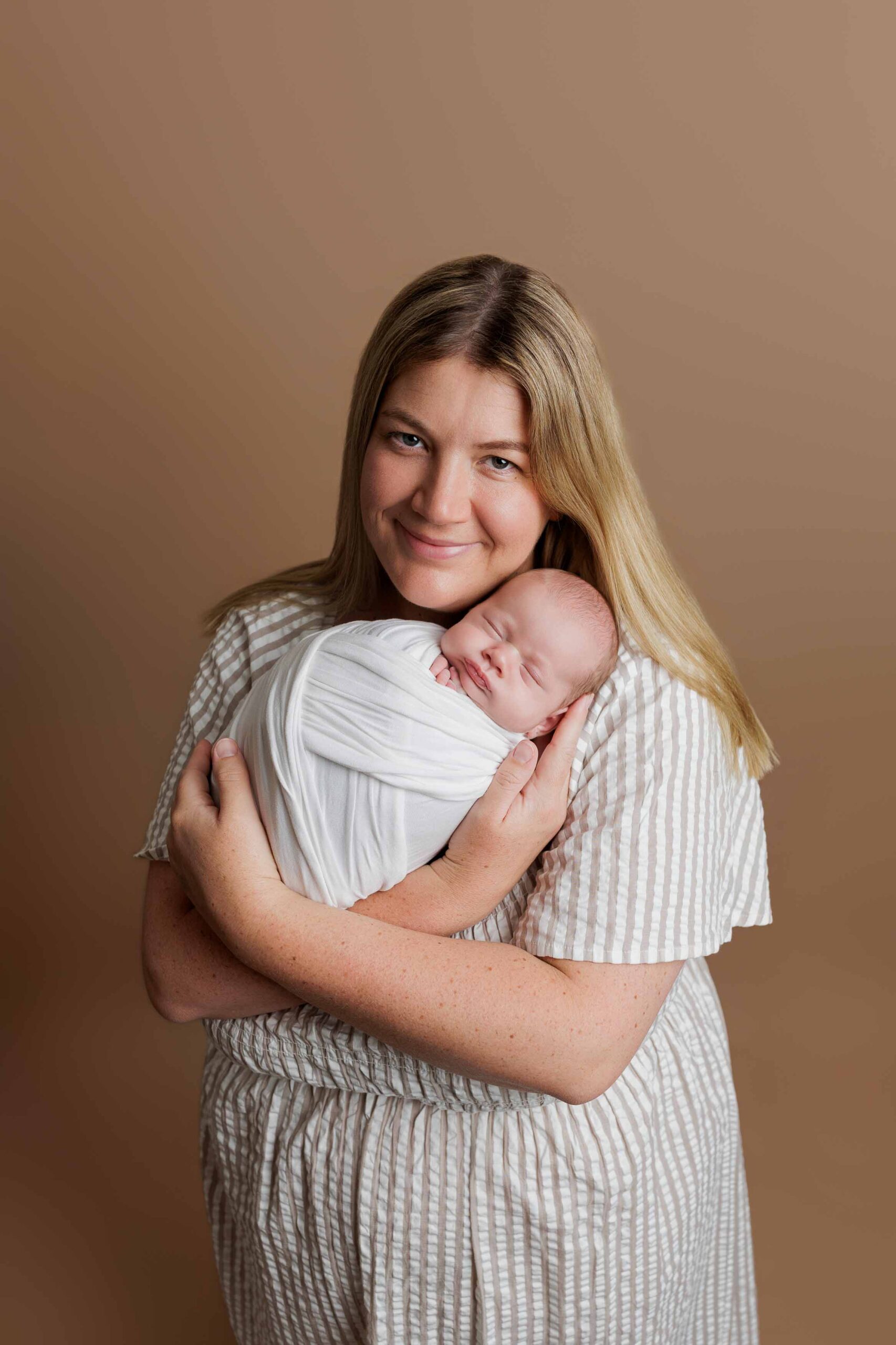 A woman with long blonde hair smiles gently whilst holding a sleeping newborn wrapped in a white blanket against a light brown background. She is wearing a light-coloured, striped dress—perfect for timeless Adelaide baby photos.