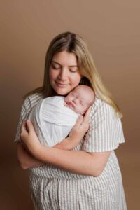 A woman with long blonde hair gently cradles a sleeping newborn wrapped in a white blanket. Both have closed eyes and peaceful expressions, captured in this serene Adelaide baby photoshoot against a plain brown background.