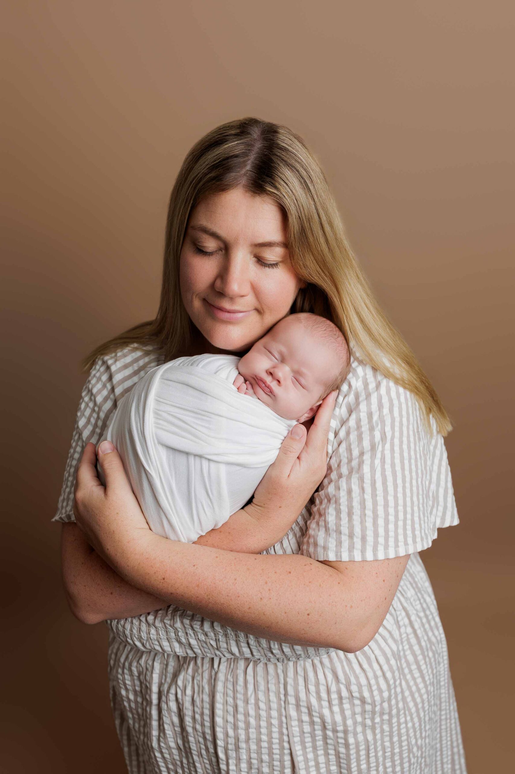 A woman with long blonde hair gently cradles a sleeping newborn wrapped in a white blanket. Both have closed eyes and peaceful expressions, captured in this serene Adelaide baby photoshoot against a plain brown background.