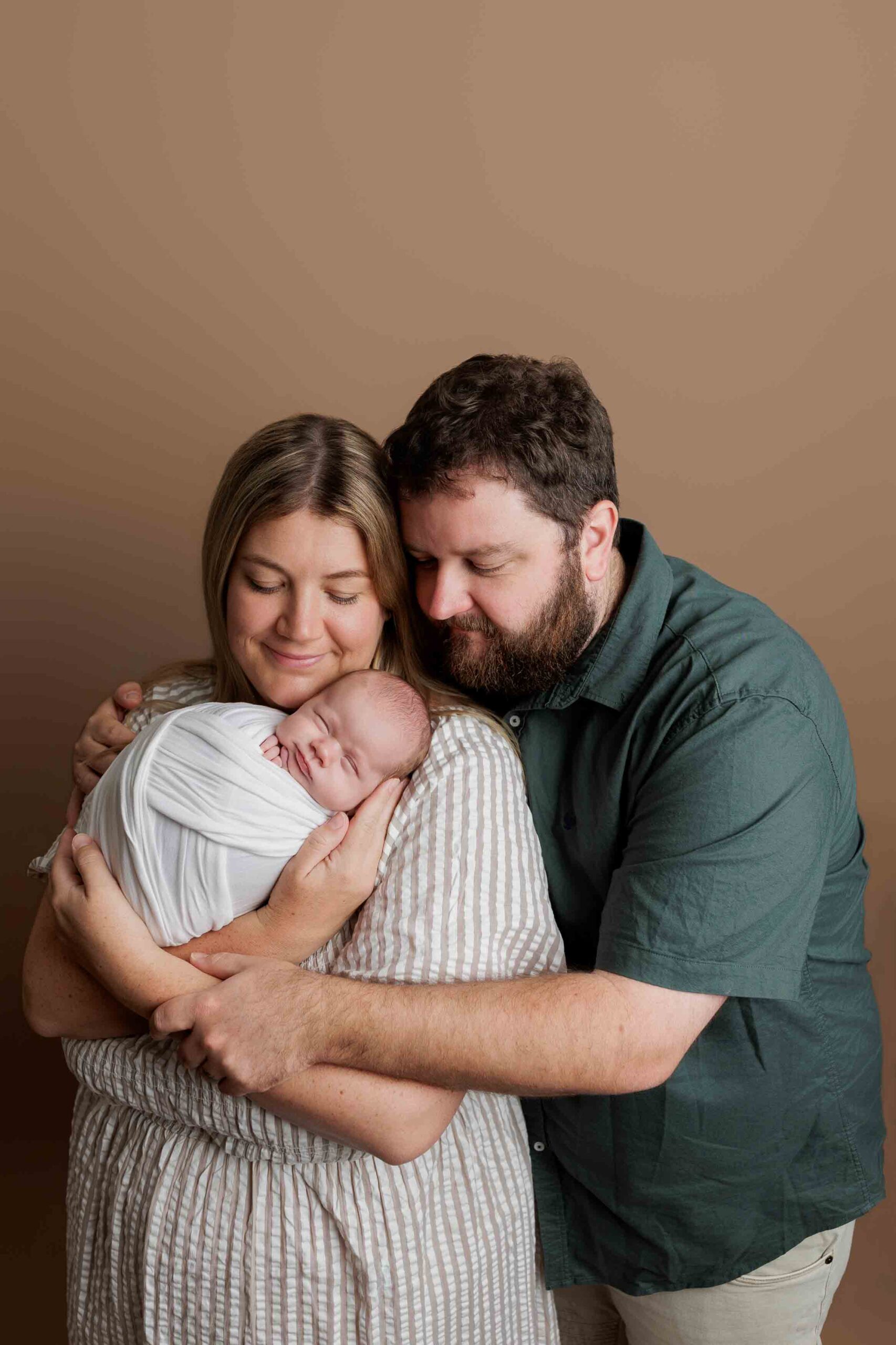 A woman and man lovingly embrace each other whilst holding a sleeping newborn wrapped in a white blanket, capturing the warmth of Adelaide baby photos against a plain brown background.