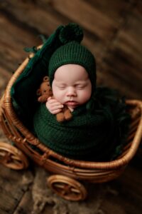 A newborn baby wrapped in a dark green knitted blanket and hat sleeps in a wicker pram, holding a small brown teddy bear. This charming scene, perfect for Adelaide baby photos, features a warm wooden floor background.