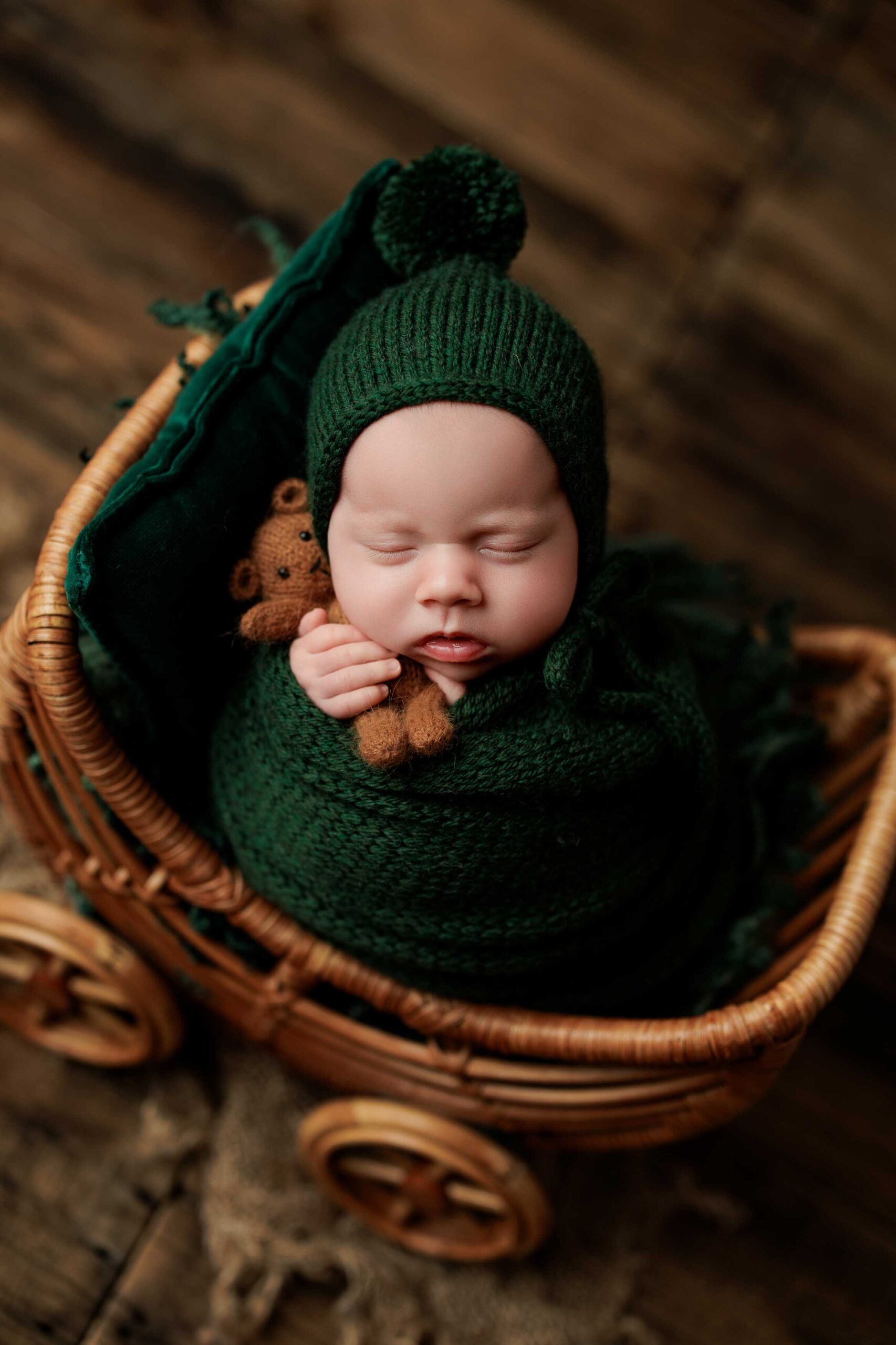 A newborn baby wrapped in a dark green knitted blanket and hat sleeps in a wicker pram, holding a small brown teddy bear. This charming scene, perfect for Adelaide baby photos, features a warm wooden floor background.