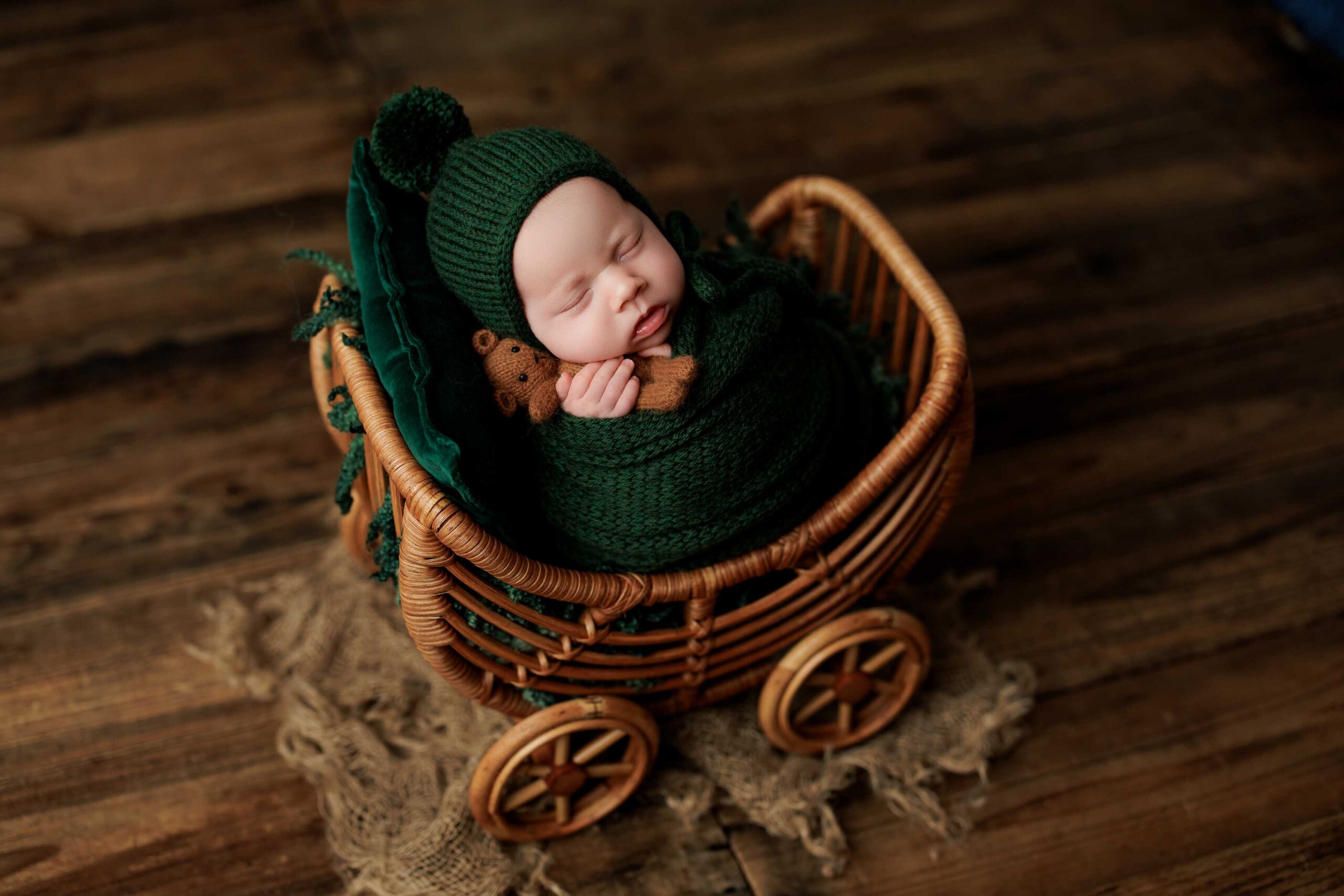 A newborn baby, wrapped in a dark green knitted blanket and hat, sleeps peacefully holding a small brown teddy bear in a wicker pram on a rustic wooden floor—perfect for Adelaide baby photos.